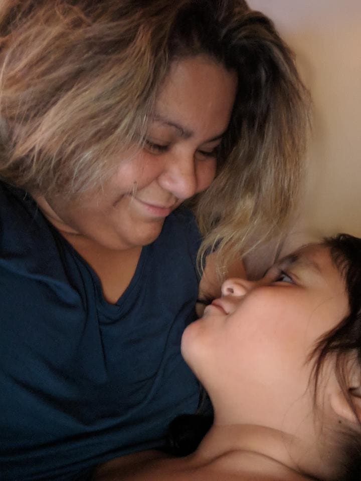 Une femme aux cheveux châtains clairs, vêtue d'un t-shirt bleu, sourit en regardant avec tendresse un jeune enfant allongé à côté d'elle. L'enfant lève les yeux vers elle, créant ainsi un moment chaleureux et plein d'affection.