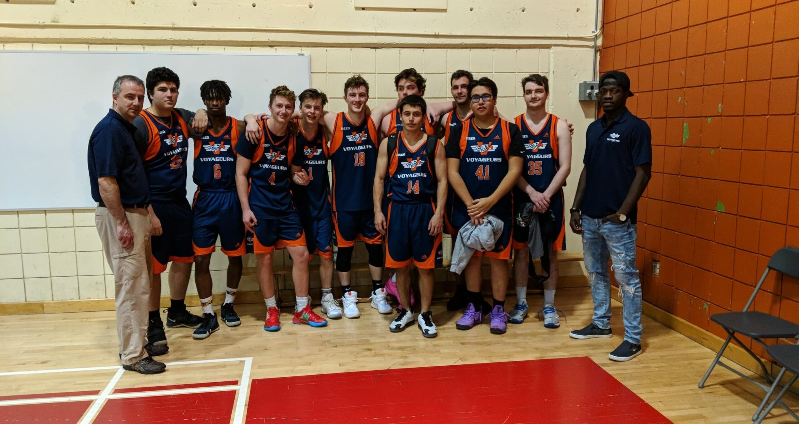 Une équipe de basket-ball, vêtue de maillots bleus et orange, pose ensemble à l'intérieur avec deux entraîneurs, debout de chaque côté, devant un tableau blanc et un mur orange.