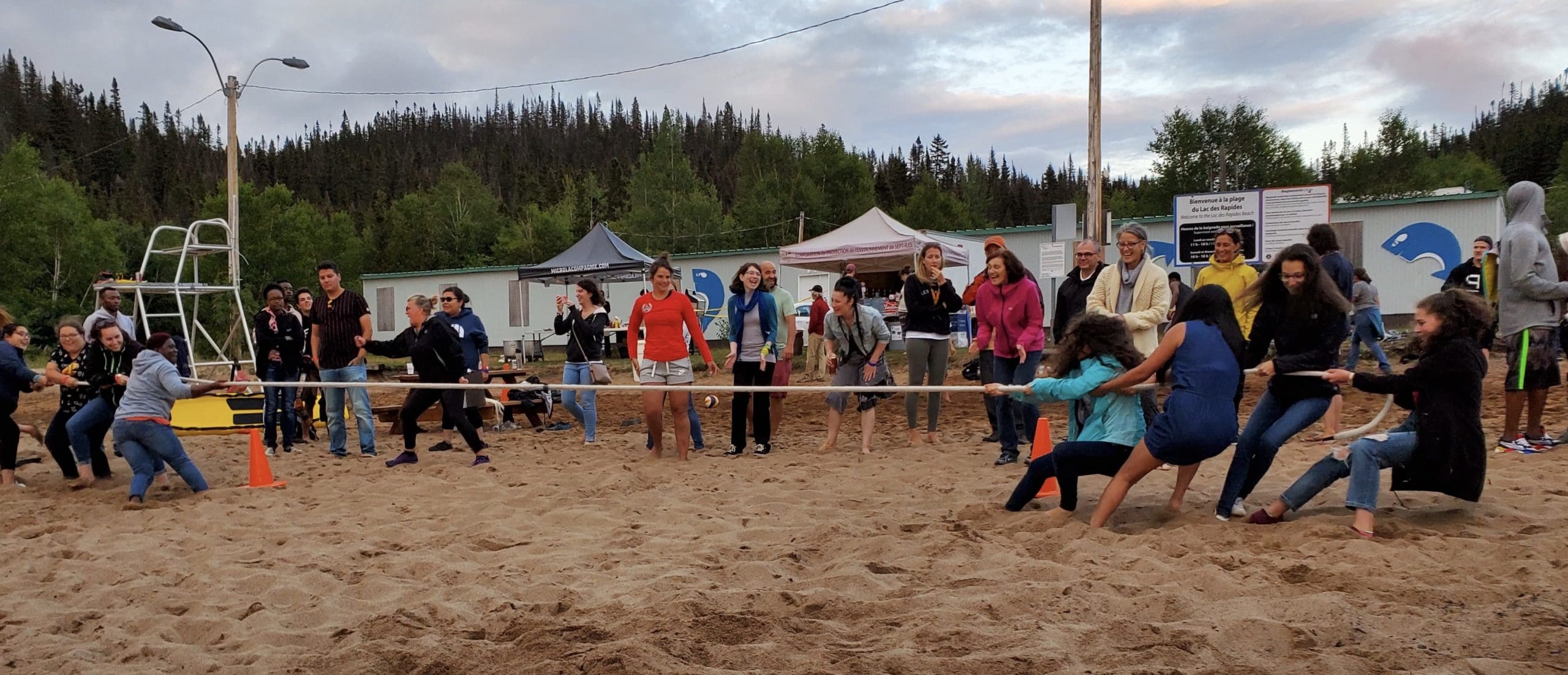 Des gens jouent au tir à la corde sur une plage de sable, encouragés par la foule. On aperçoit des tentes et des arbres à l'arrière-plan ; l'événement semble animé et très disputé.