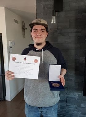 Un jeune homme, coiffé d'une casquette et vêtu d'un sweat à capuche, sourit, tenant d'une main un certificat et de l'autre un coffret à médailles ouvert ; il se tient à l'intérieur, près d'un mur sombre.