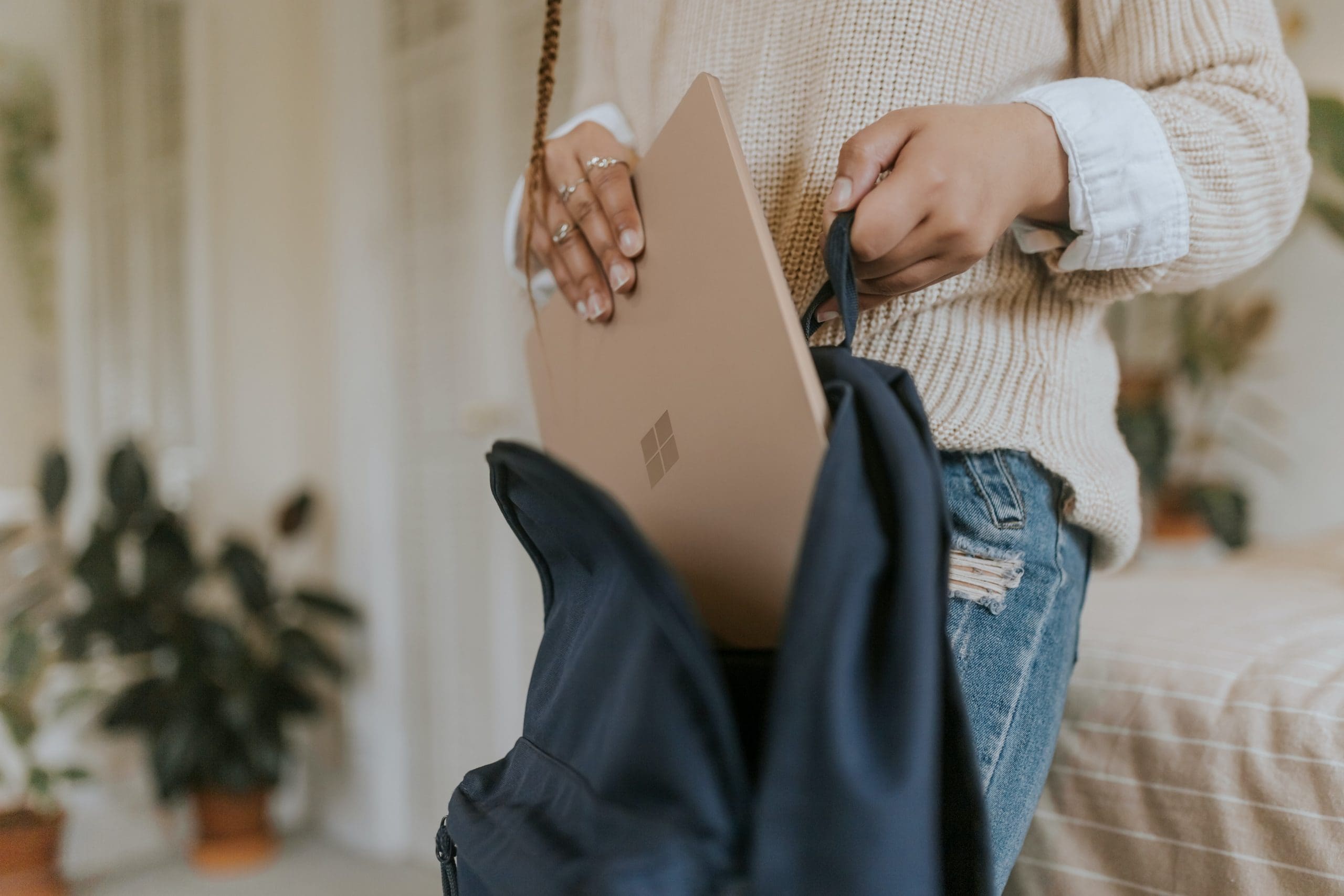 Une personne vêtue d'un pull beige et d'un jean range un ordinateur portable couleur beige clair dans un sac à dos noir, dans une pièce chaleureuse et remplie de plantes.