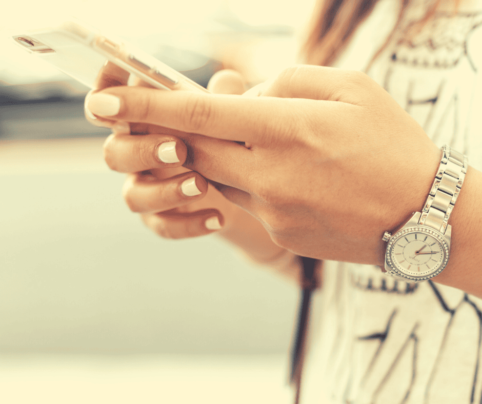 Gros plan sur les mains d'une personne tenant un smartphone. Cette personne a les ongles vernis d'une couleur claire et porte une montre-bracelet argentée. L'arrière-plan est flou.