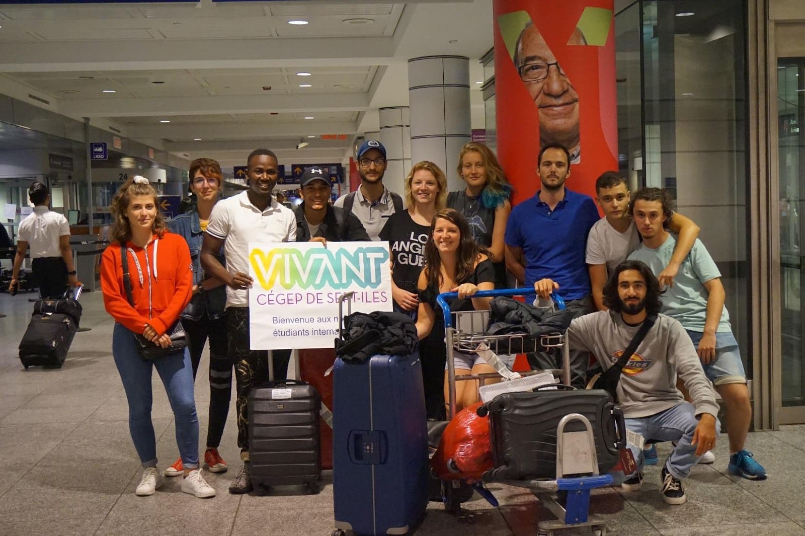 Un groupe de 13 personnes, bagages à la main, pose dans un aéroport en tenant une pancarte sur laquelle on peut lire « VIVANT CÉGEP DE SEPT-ÎLES ». Certaines sourient, et deux chariots à bagages chargés de valises sont garés devant elles.