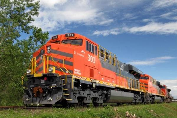 Une locomotive de train de marchandises orange vif, portant le numéro « 301 » à l'avant et sur le côté, roule sur des voies ferrées sous un ciel bleu parsemé de nuages, avec de la verdure à proximité.