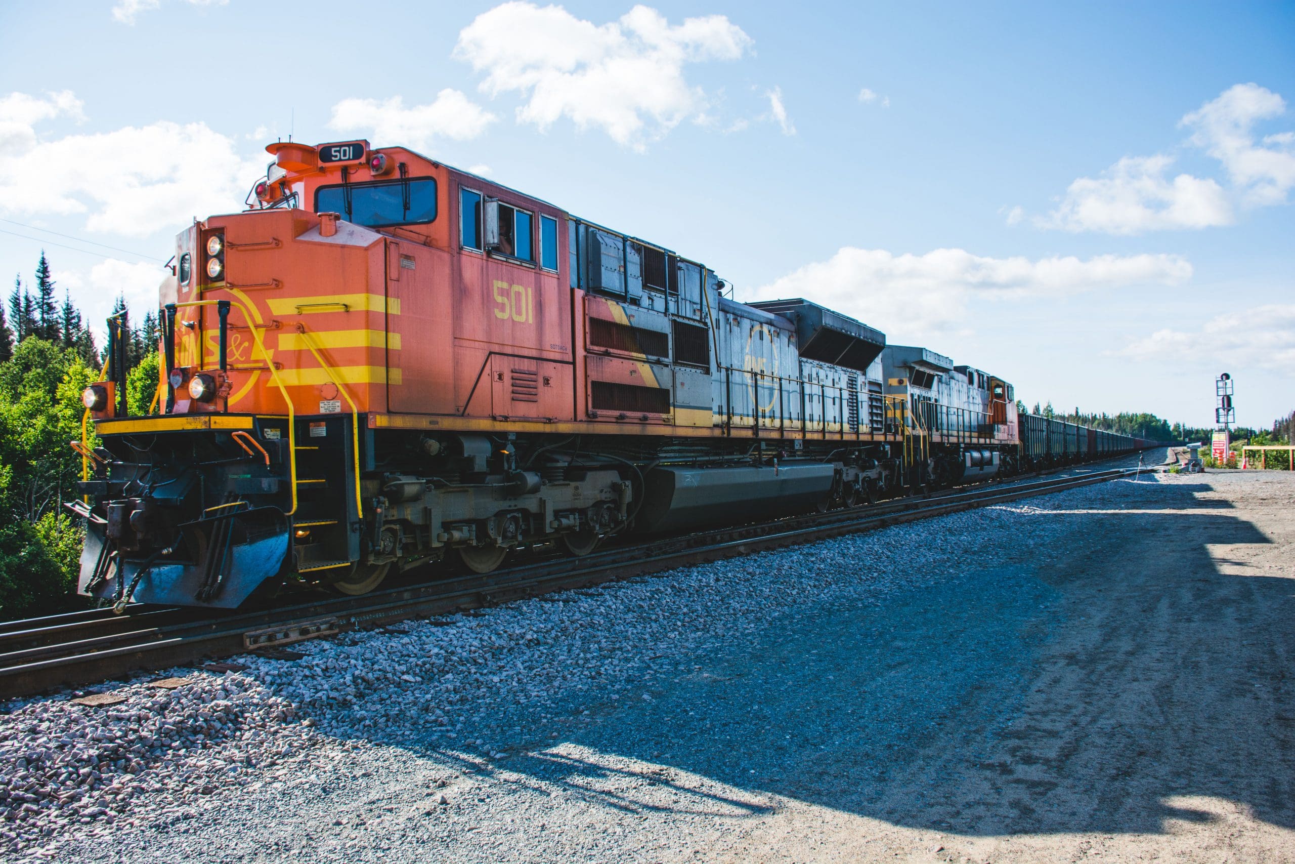 Une locomotive orange et jaune, portant le numéro 501, tracte un long train de marchandises sur des voies ferrées, avec des arbres en arrière-plan sous un ciel bleu parsemé de nuages.
