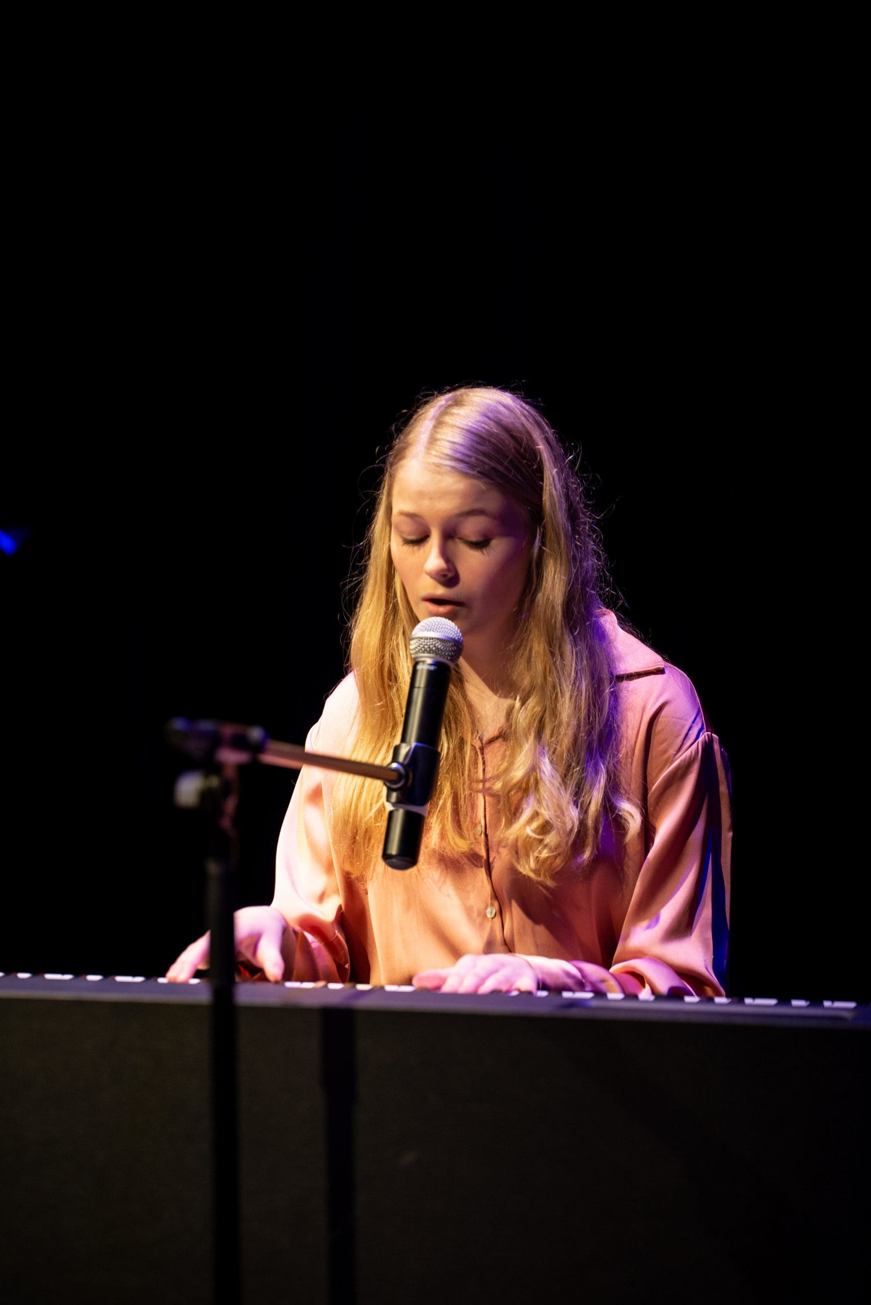 Une jeune femme aux longs cheveux blonds joue du clavier et chante au micro sur scène, éclairée par un projecteur sur fond sombre.
