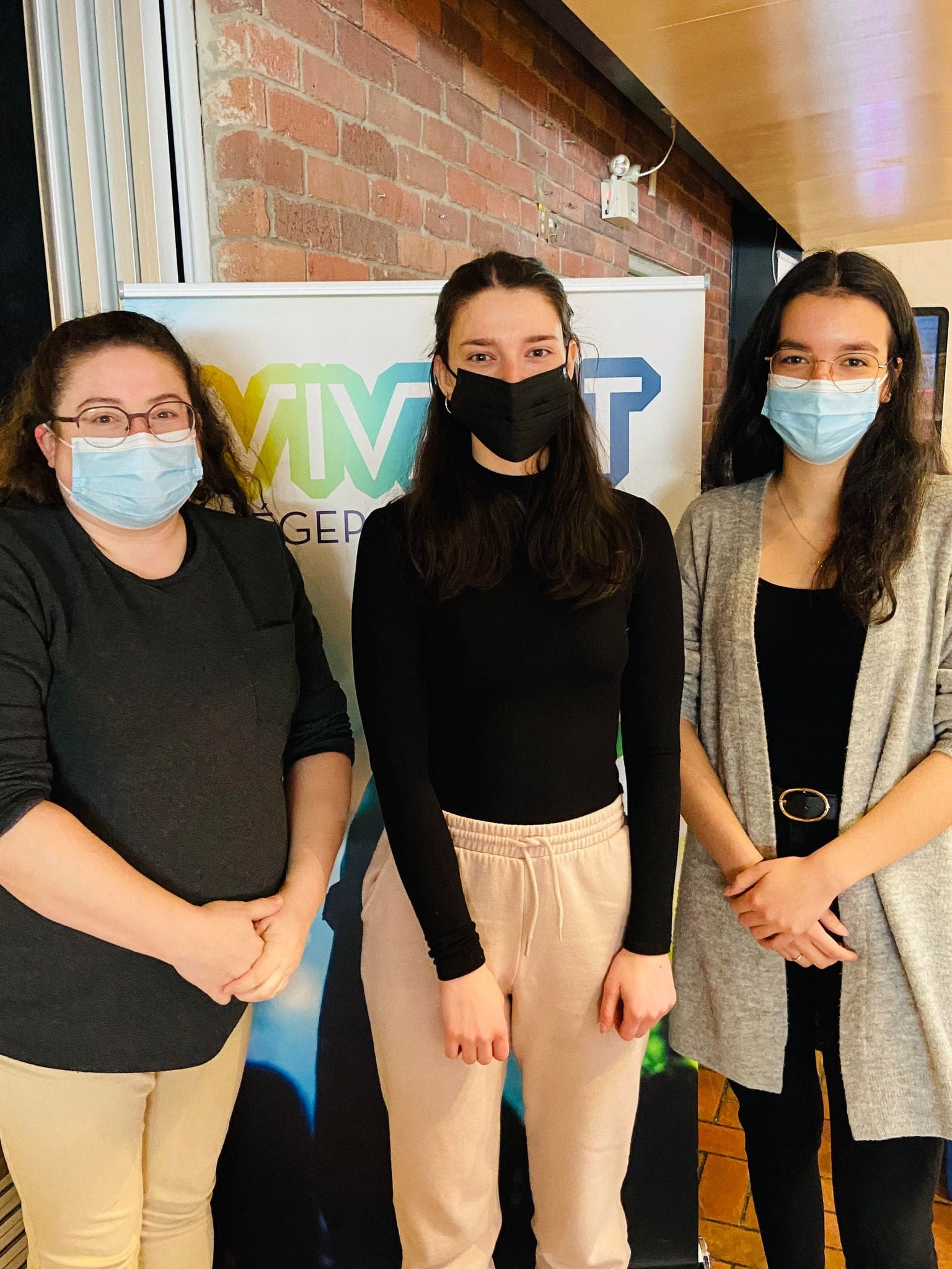 Trois femmes portant des masques se tiennent à l'intérieur, devant un mur de briques et une pancarte sur laquelle figurent des inscriptions en vert et en bleu. Elles regardent l'appareil photo et prennent la pose ensemble.