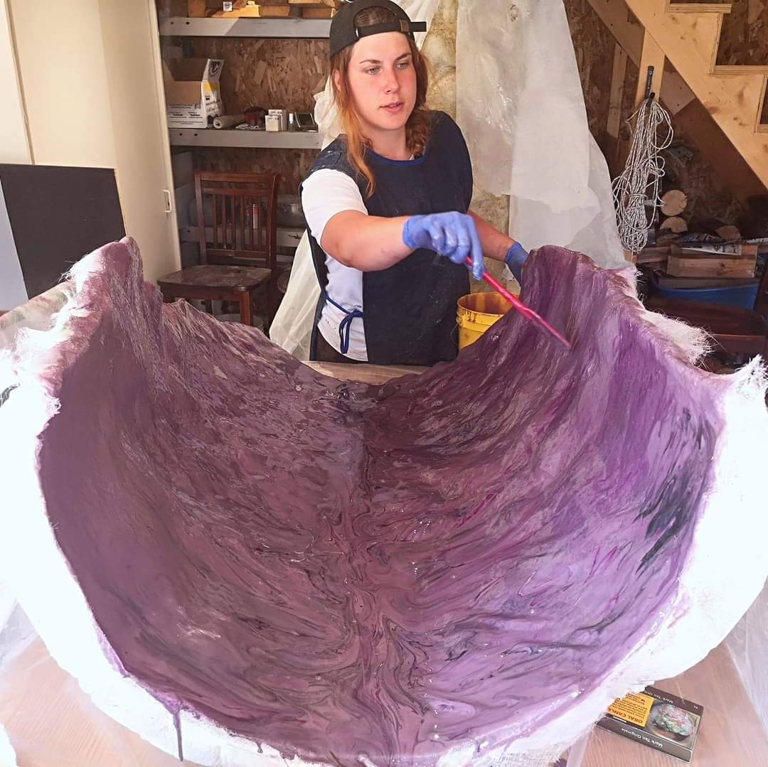 Dans un atelier, une personne portant des gants et une casquette à l'envers peint l'intérieur d'une grande structure violette et incurvée à l'aide d'un pinceau.