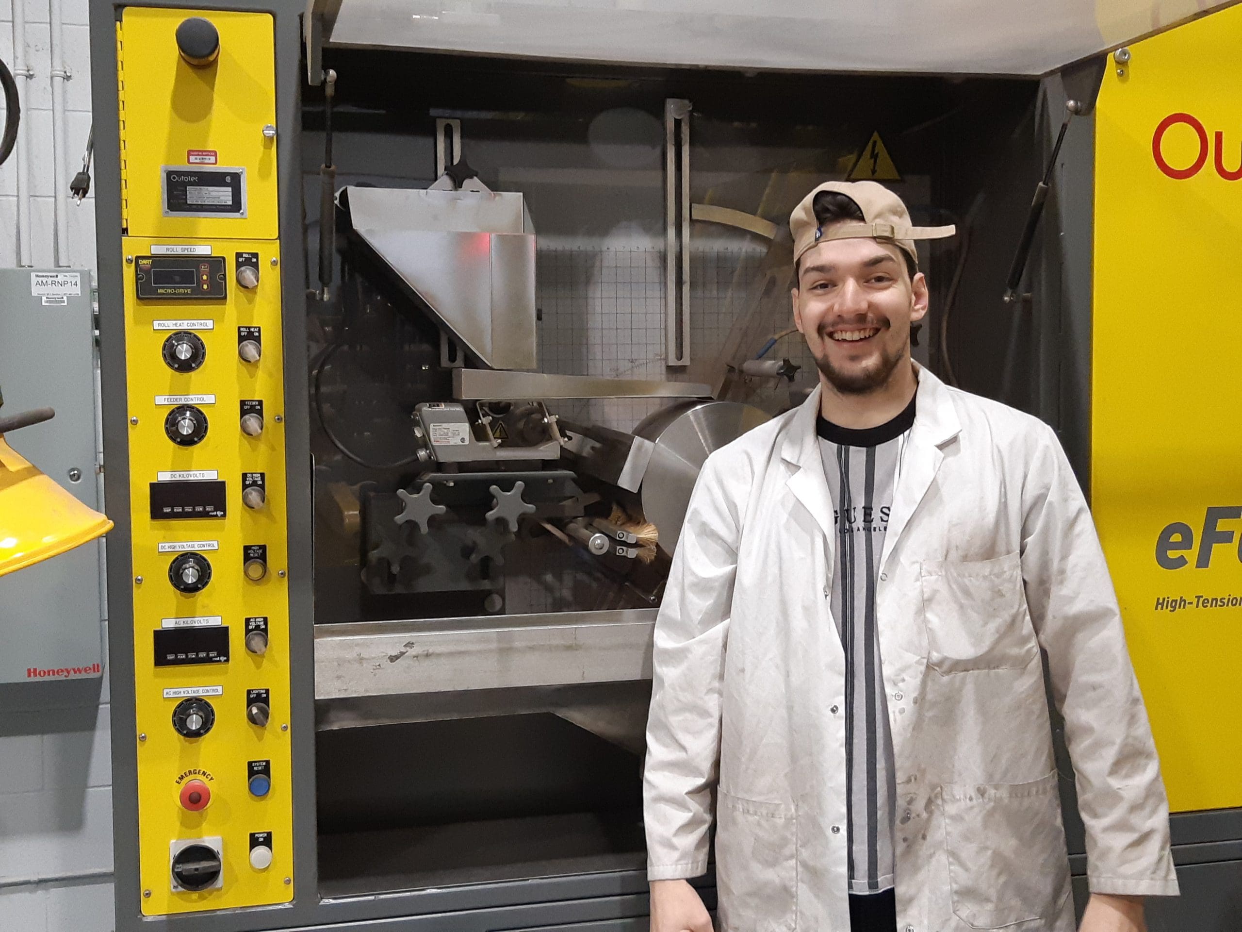 Une personne souriante, vêtue d'une blouse blanche et d'une charlotte, se tient devant une grande machine industrielle dotée de panneaux jaunes et de divers boutons de commande.