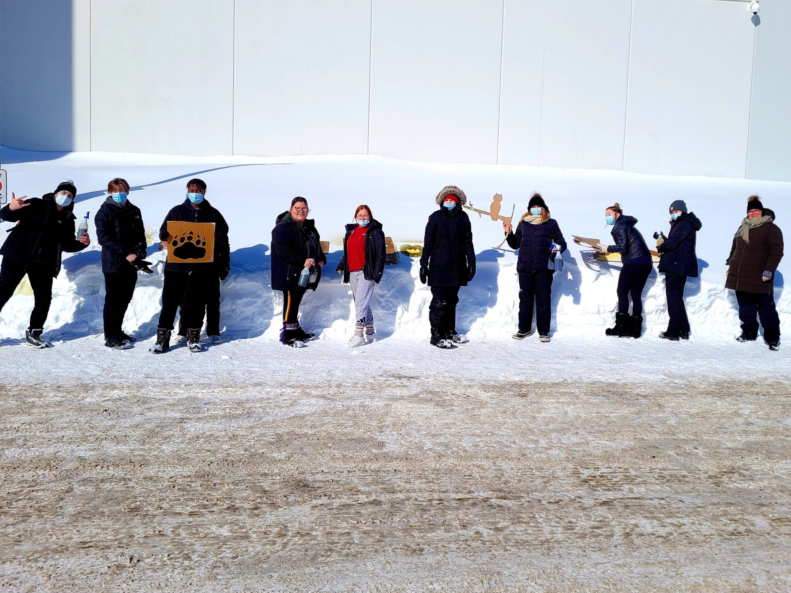Neuf personnes se tiennent dans un espace enneigé près d'un mur, vêtues de tenues d'hiver et portant des masques. Certaines tiennent des objets, comme une pancarte représentant une patte d'ours et des récipients contenant de la nourriture. La lumière vive du soleil projette des ombres marquées sur la neige.