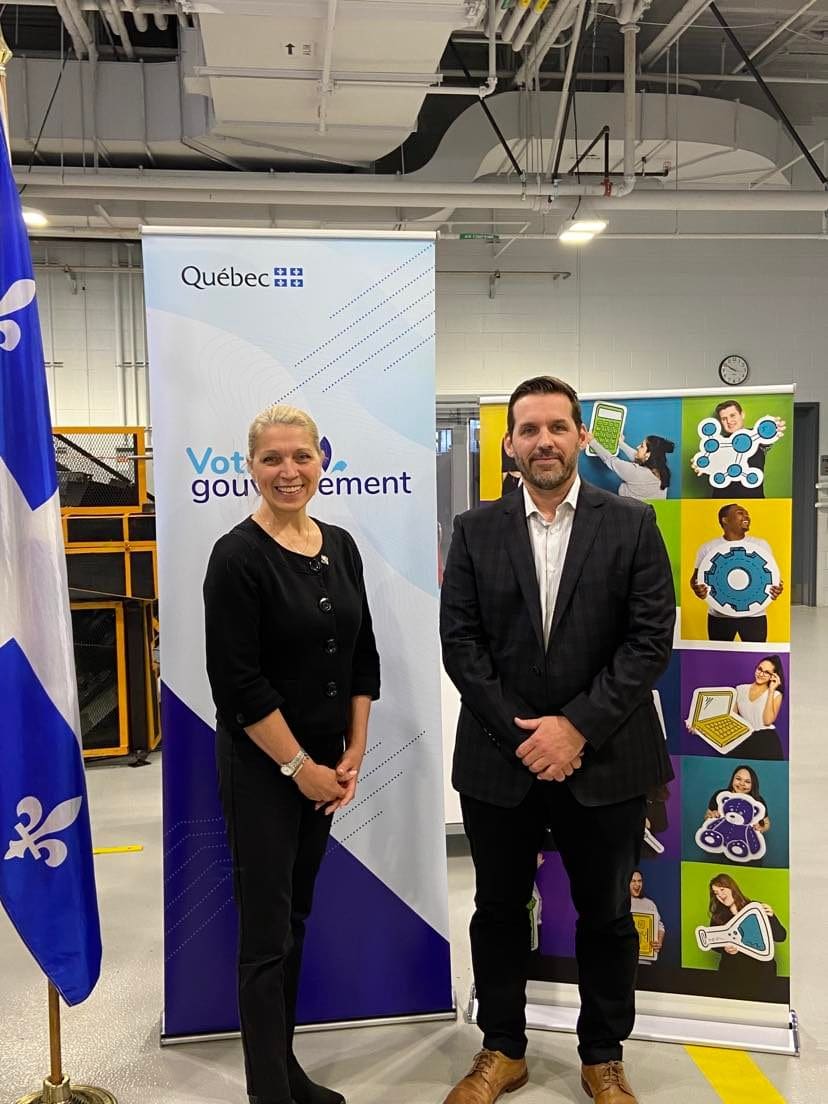 Deux personnes se tiennent à l'intérieur, entre des banderoles arborant les logos du gouvernement du Québec et des images colorées. Un drapeau du Québec est visible à gauche. Le décor semble être un environnement industriel ou un lieu de travail.