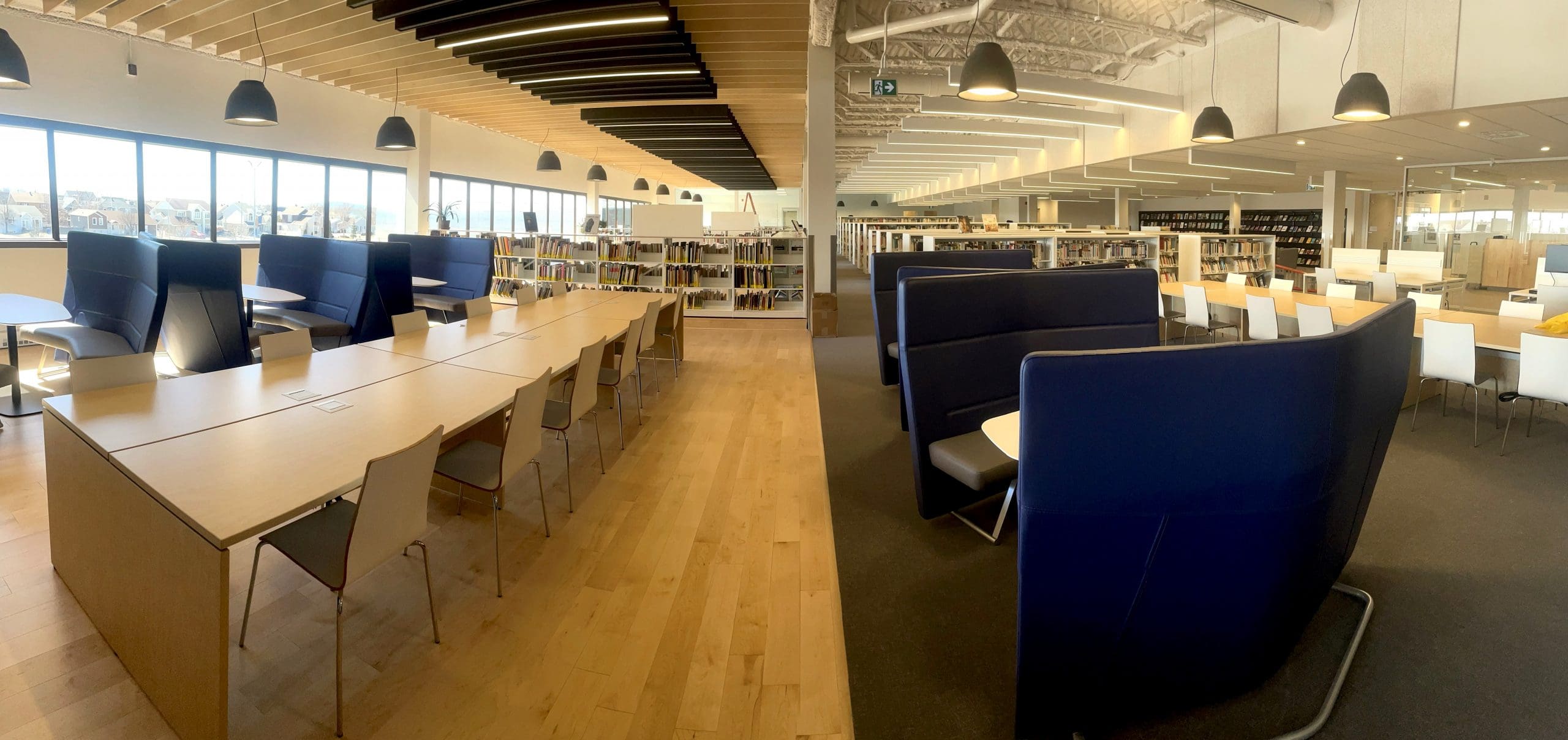 Une bibliothèque spacieuse et moderne, dotée de tables en bois, de sièges rembourrés bleus, d'étagères, de grandes fenêtres et de suspensions lumineuses, créant ainsi un espace d'étude lumineux et accueillant.