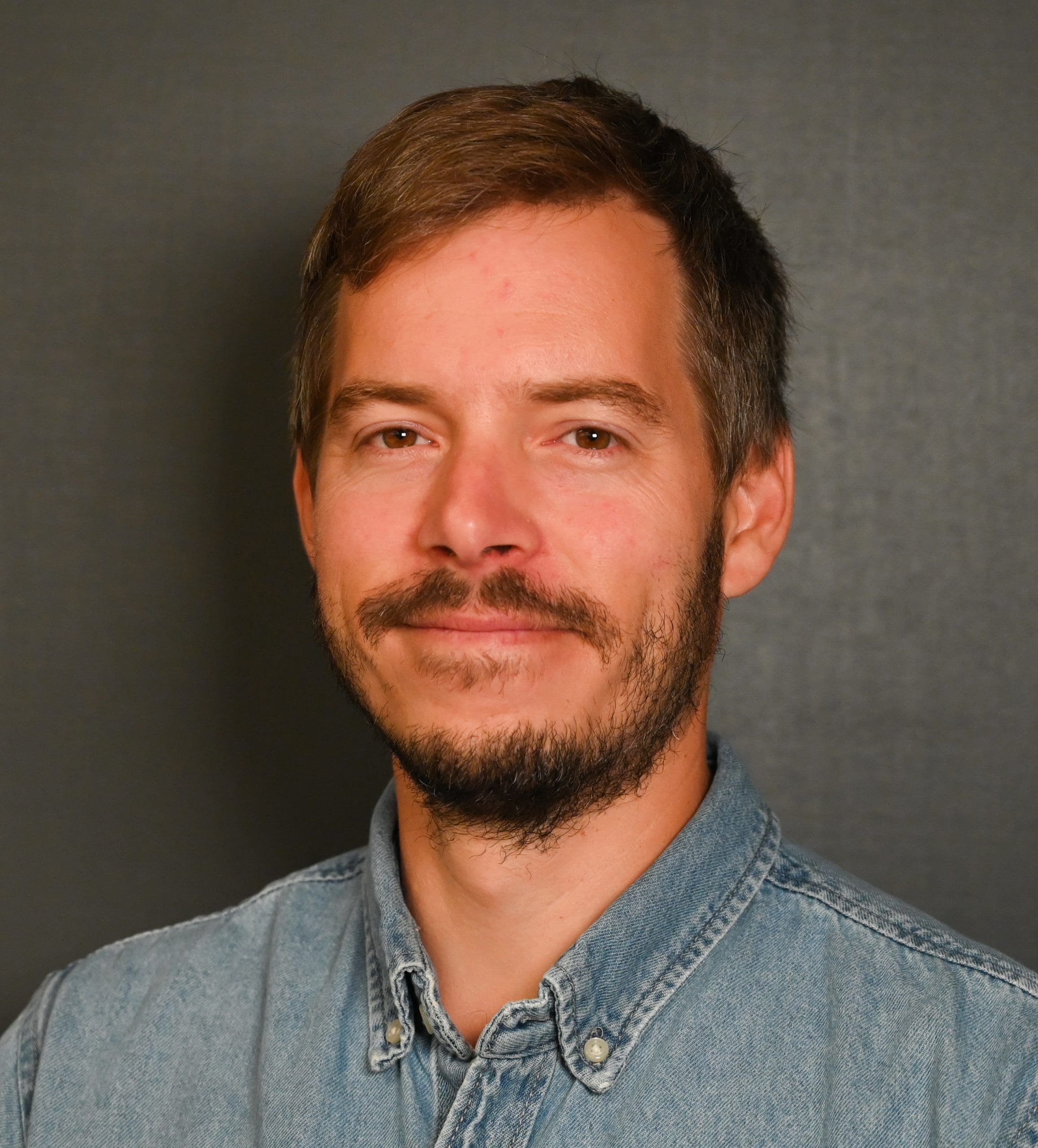 Un homme aux cheveux bruns courts et à la barbe, vêtu d'une chemise en jean bleu clair, sourit doucement devant un fond sombre et uni.