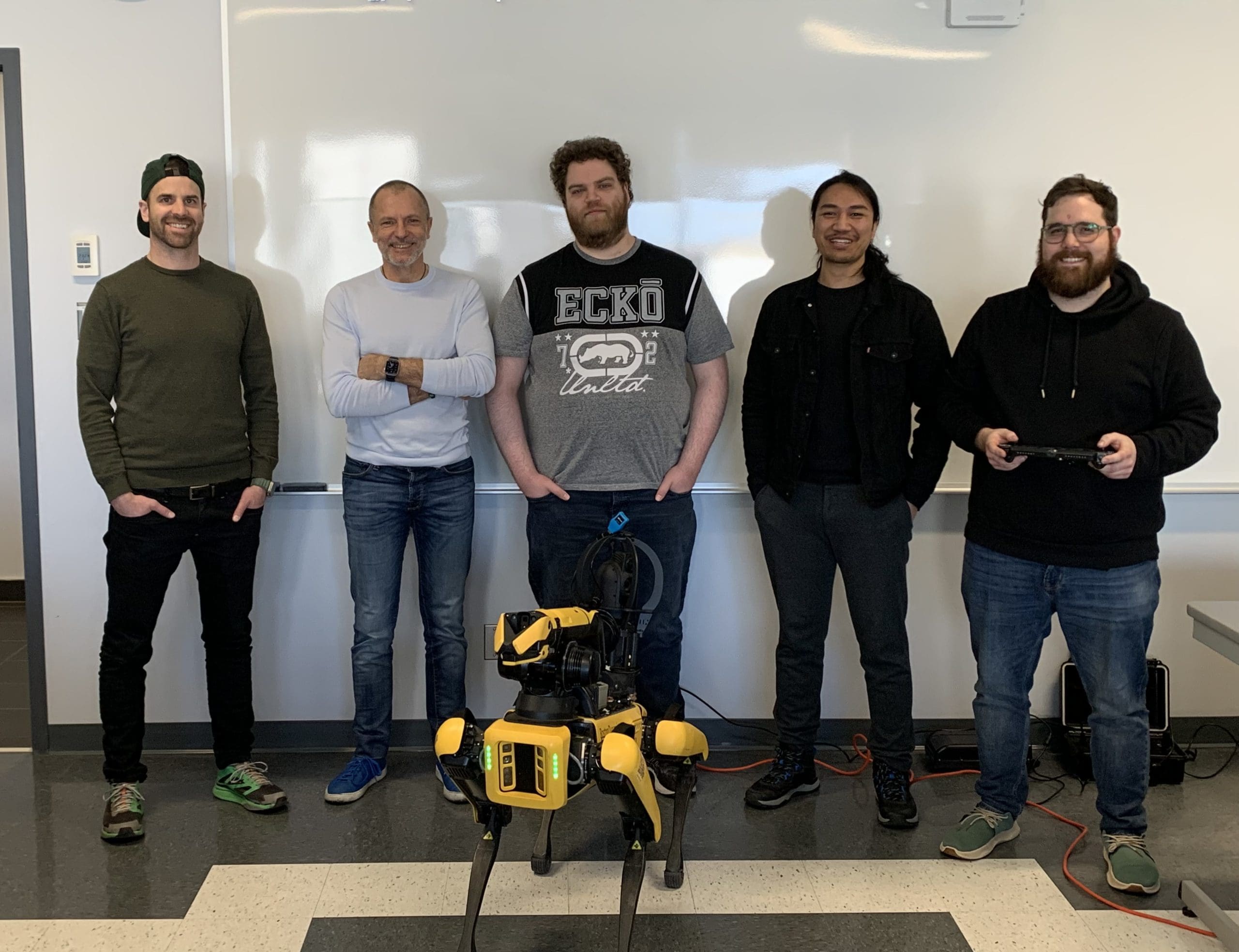 Cinq hommes se tiennent à l'intérieur devant un tableau blanc, souriants, avec un chien robot jaune placé devant eux. Une personne, à droite, tient une manette.