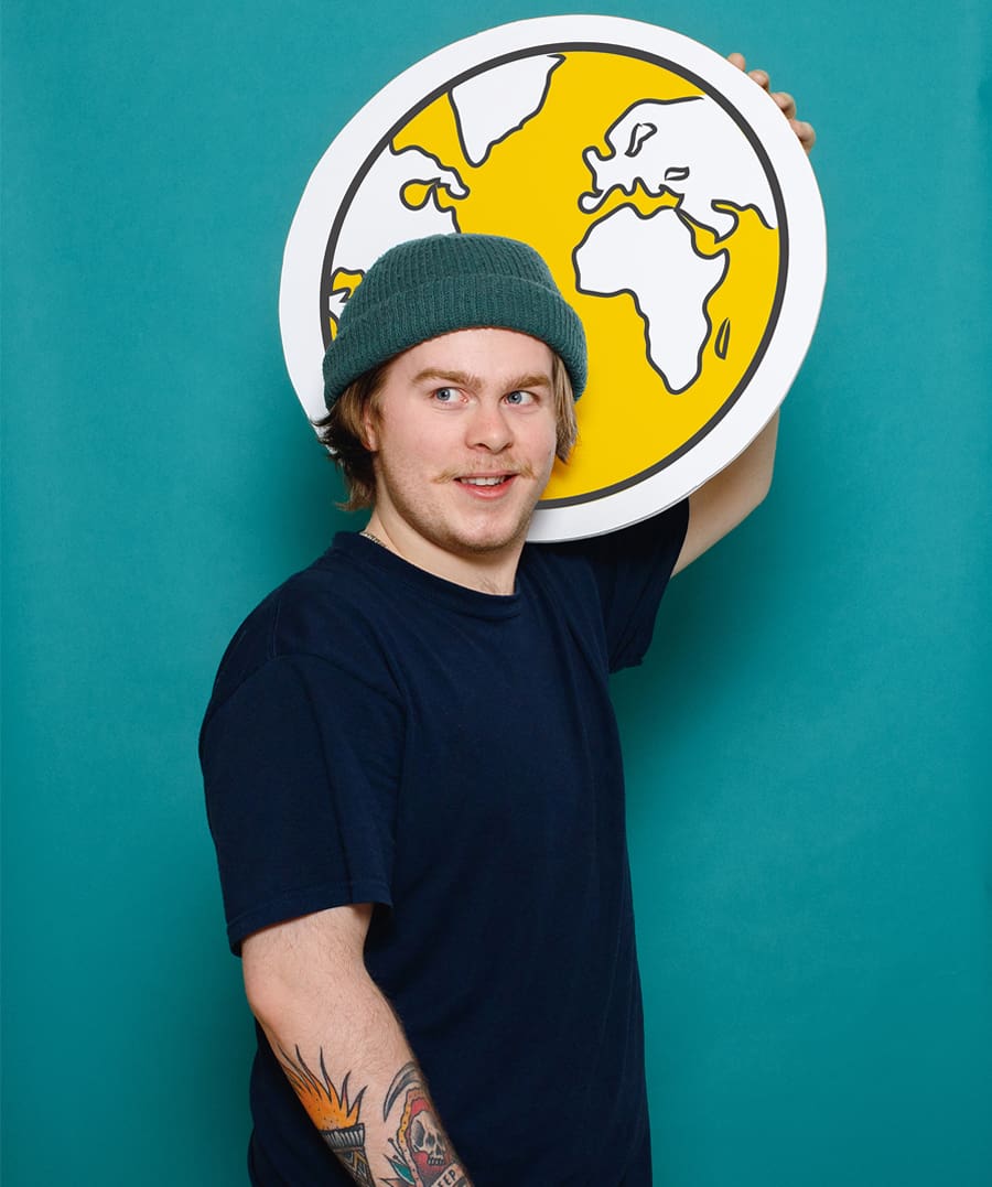 Une personne portant un bonnet vert et un t-shirt foncé tient une pancarte ronde sur laquelle figure une carte du monde jaune, se détachant sur un fond bleu sarcelle. On aperçoit un tatouage sur son avant-bras.