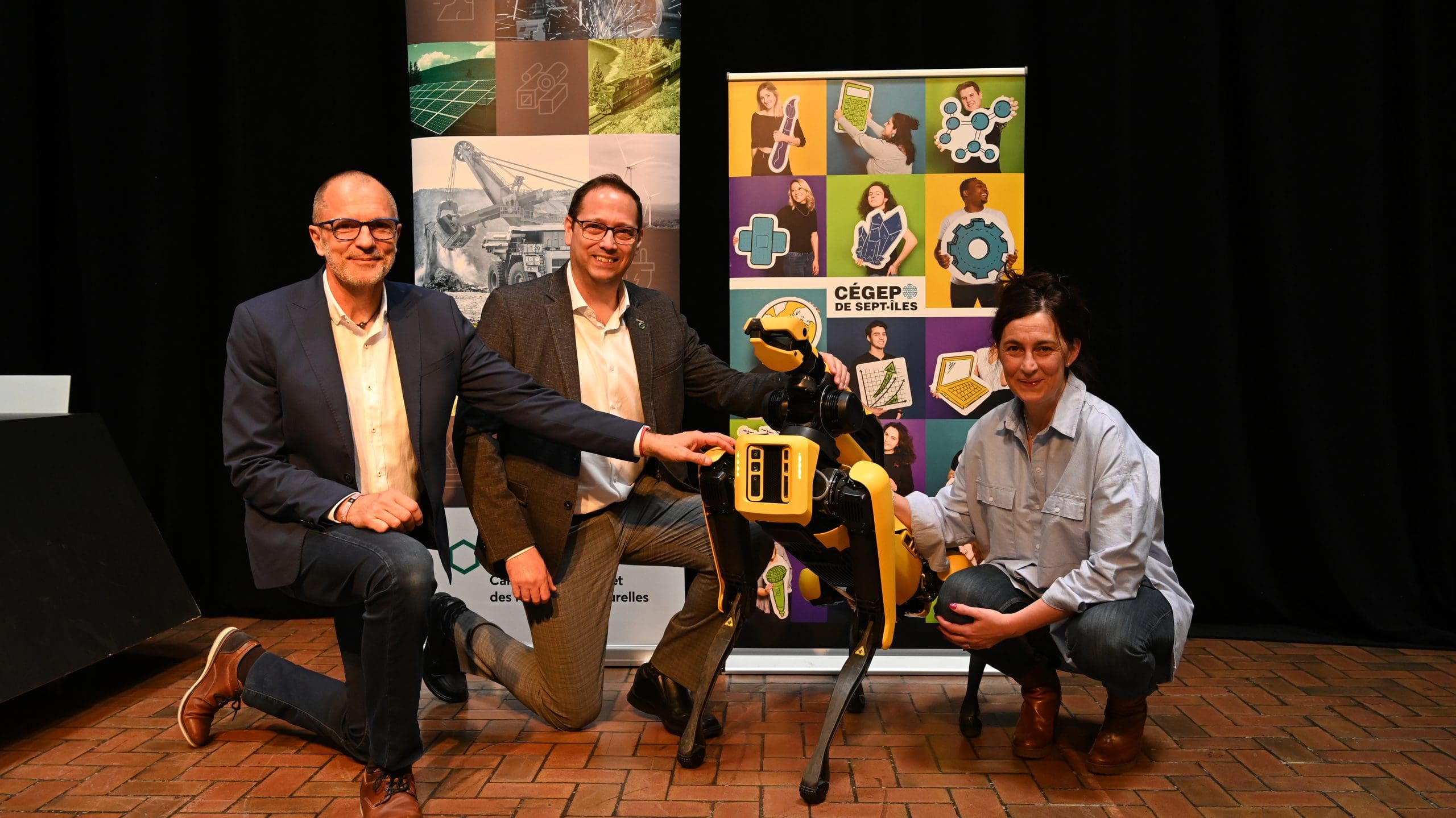 Trois personnes sont agenouillées à côté d'un chien robot jaune, devant des banderoles colorées, et sourient à la caméra sur un sol en briques, à l'intérieur d'un bâtiment.