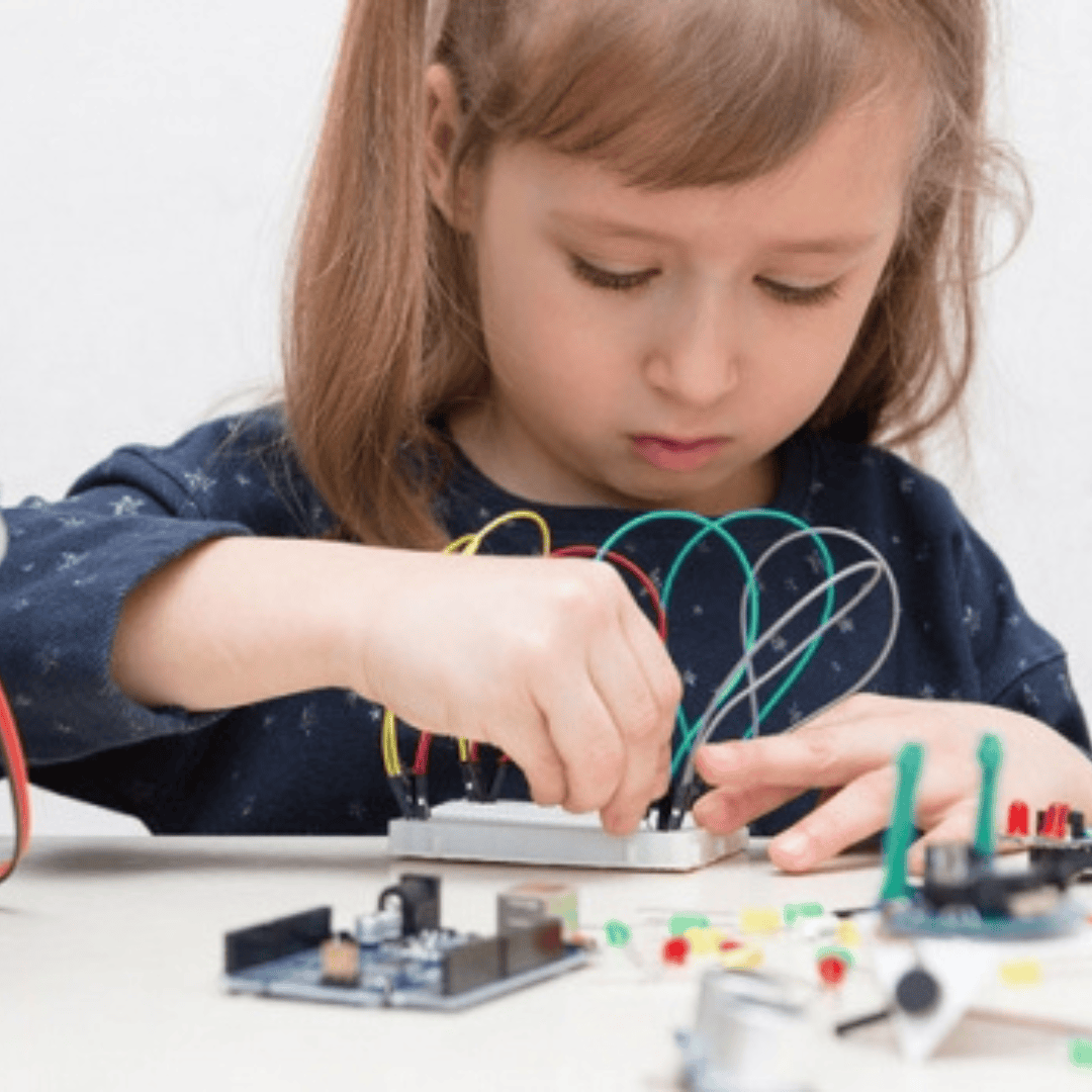 Une jeune fille est concentrée sur son travail : elle assemble des fils colorés et des composants électroniques sur un petit circuit imprimé, assise à une table.