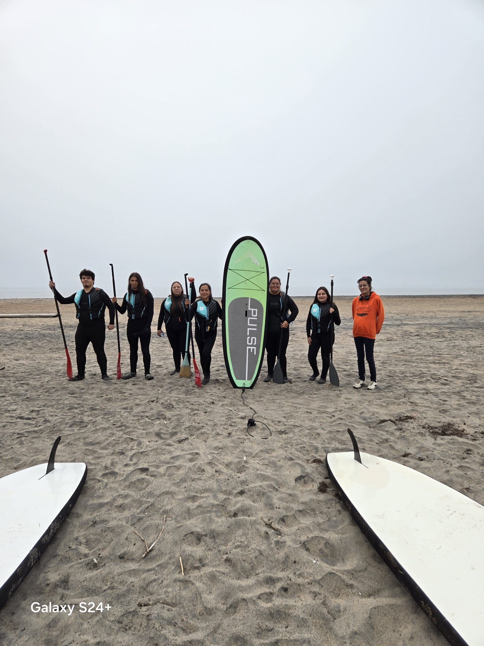 Huit personnes en combinaison de plongée se tiennent sur une plage, une pagaie à la main, et prennent la pose autour d'une planche de paddle verte et blanche posée à la verticale dans le sable. Deux autres planches de paddle sont visibles au premier plan. Le ciel est couvert.
