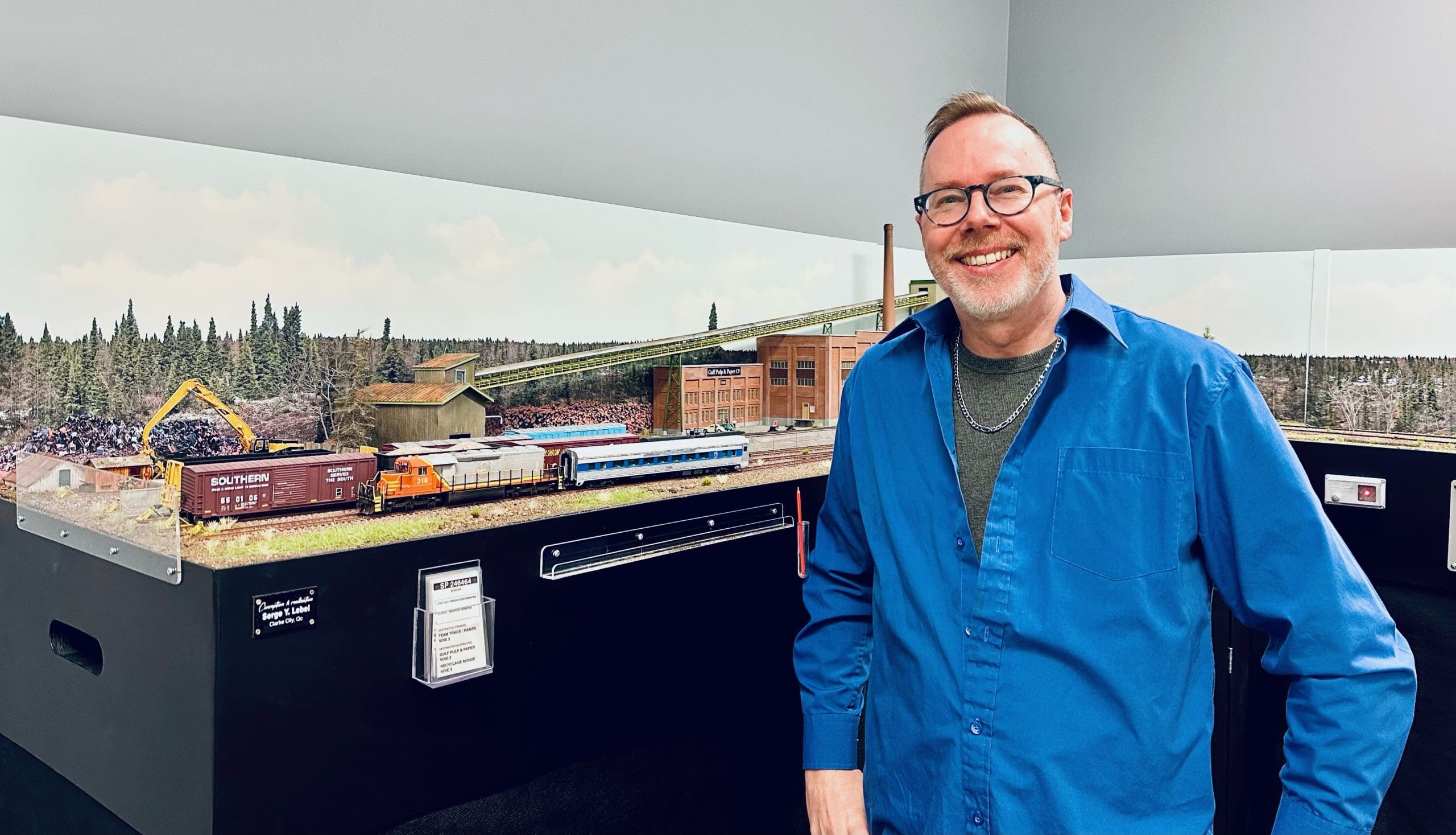 Un homme souriant, portant des lunettes et une chemise bleue, se tient à côté d'un maquette de train très détaillée, comprenant des arbres, des bâtiments et plusieurs wagons, dans une pièce aux murs gris clair.