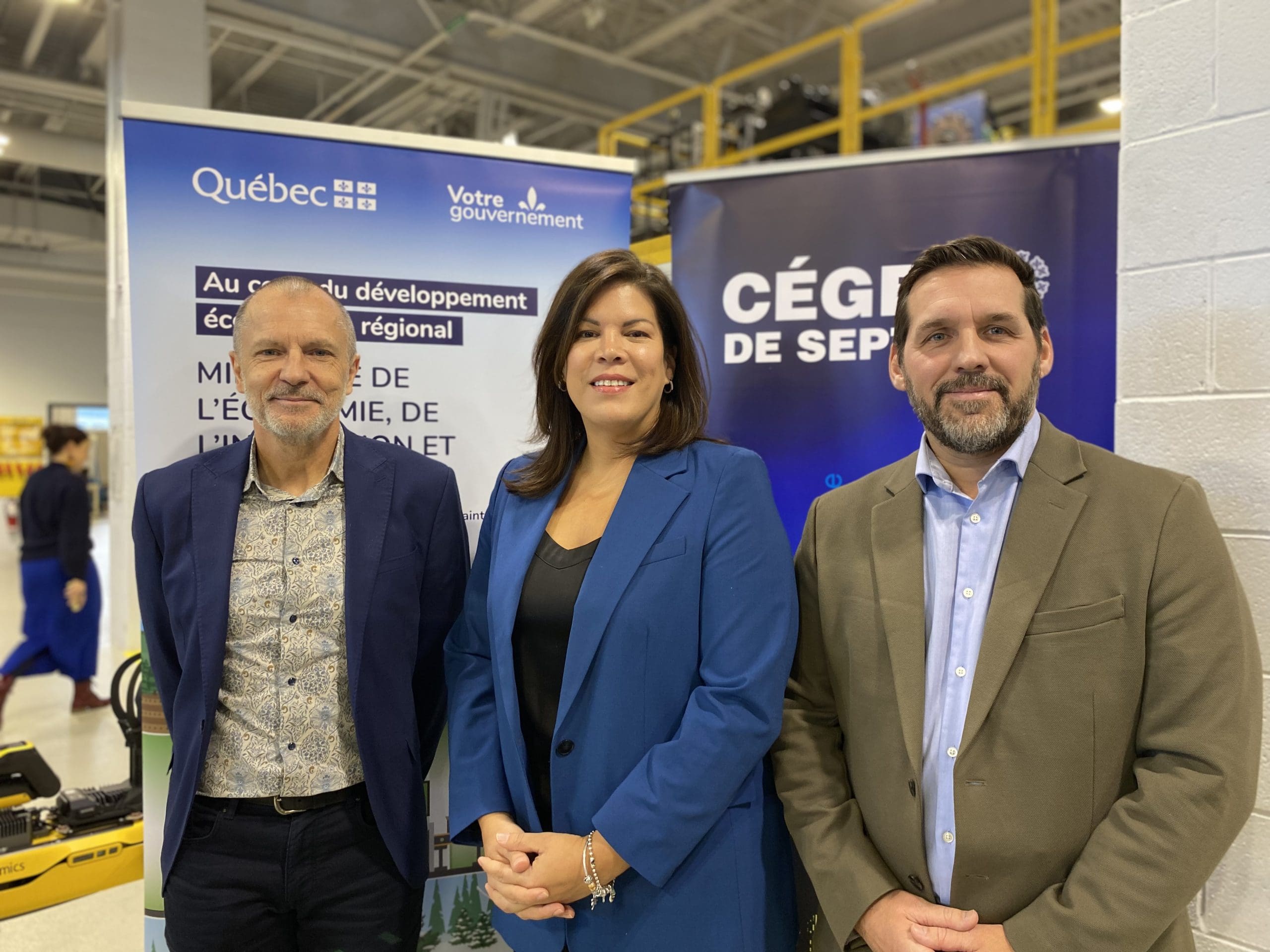 Trois personnes en tenue professionnelle se tiennent debout et sourient devant des bannières représentant le Québec et le Cégep de Sept-Îles, à l'intérieur d'un bâtiment moderne à vocation industrielle ou éducative.
