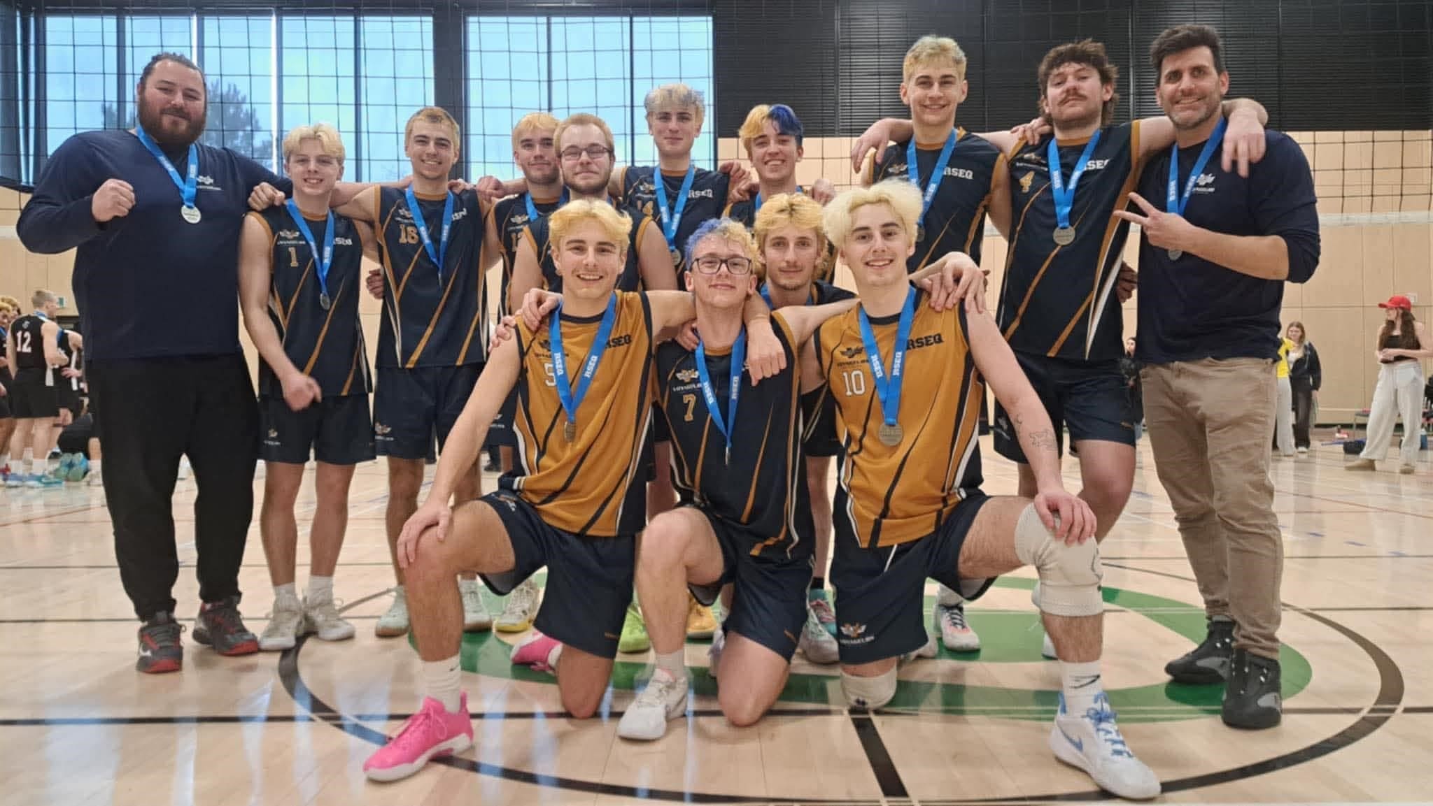 Une équipe masculine de volley-ball, arborant ses médailles, pose ensemble sur le terrain d'un gymnase. La plupart des joueurs portent des maillots bleu marine et dorés, et deux entraîneurs, vêtus de chemises bleu marine, se tiennent aux extrémités du terrain. Le groupe sourit fièrement pour la photo.