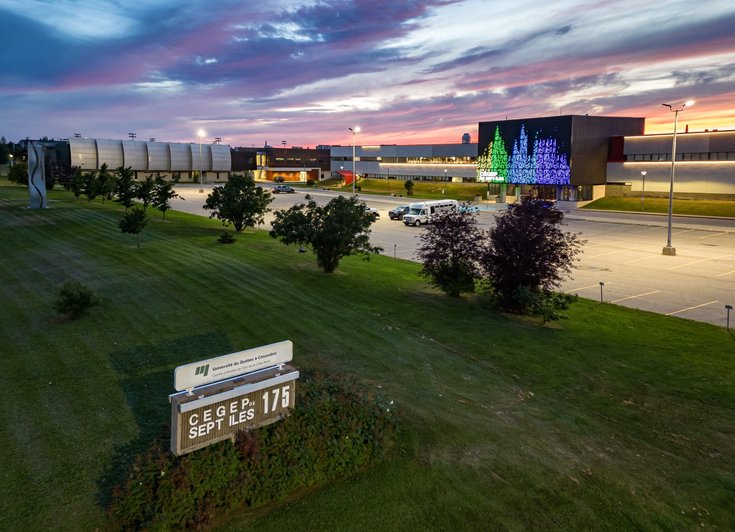 Un bâtiment moderne au coucher du soleil, avec sur sa façade des lumières colorées en forme d'arbres, un parking, une pelouse verte, des arbres et un panneau sur lequel on peut lire « CÉGEP SEPT-ÎLES 175 ».