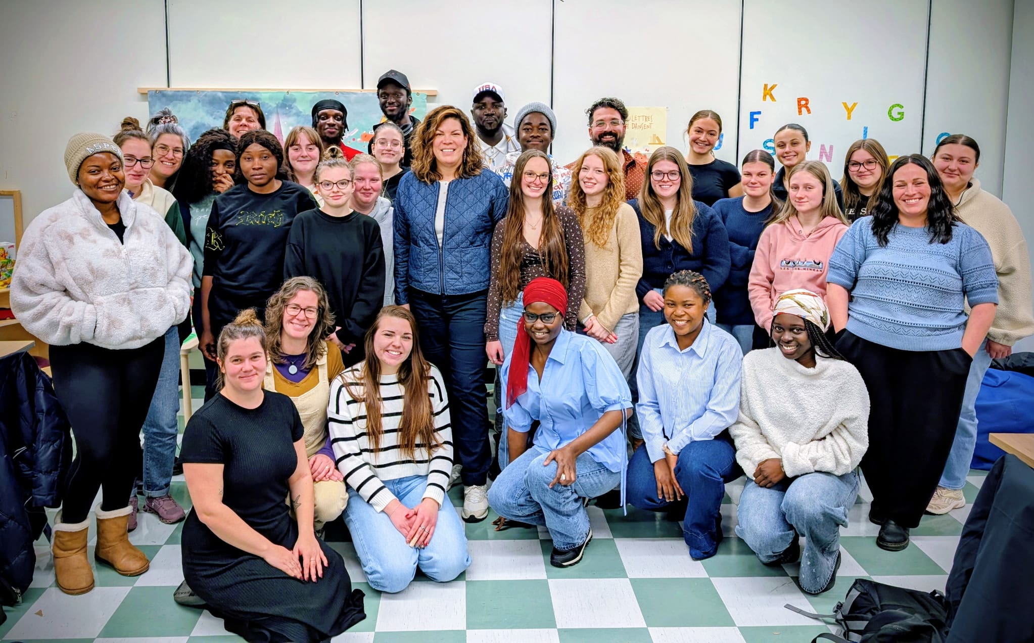 Un groupe d'une trentaine de personnes pose ensemble dans une pièce très éclairée, souriant à l'appareil photo. Certaines sont debout, d'autres à genoux. Des lettres colorées et des illustrations ornent le mur en arrière-plan.