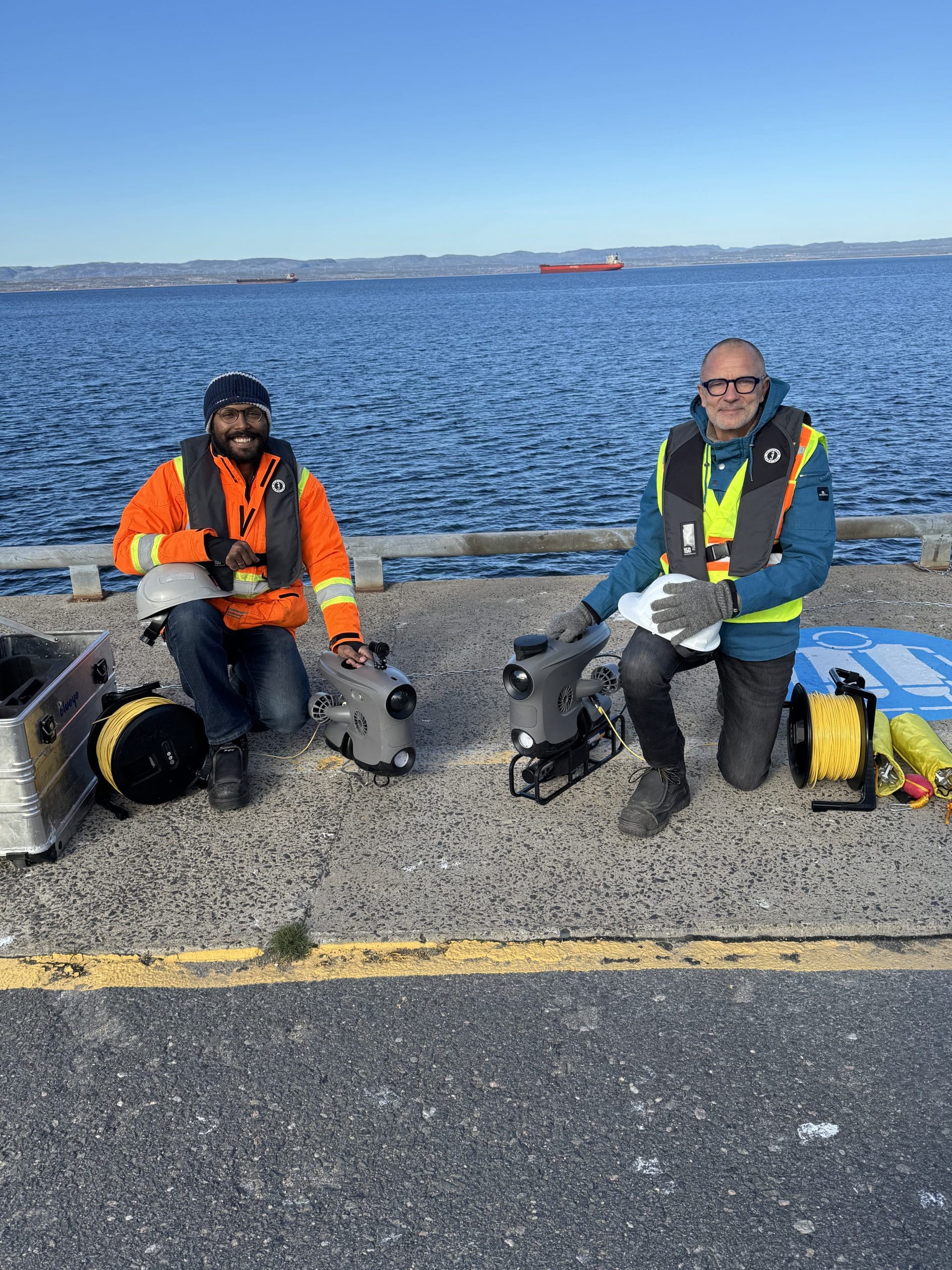 Deux personnes sont agenouillées sur un quai au bord de l'eau ; elles portent des gilets de sécurité et préparent des véhicules sous-marins télécommandés (ROV) à l'aide de câbles et de matériel disposés à côté d'elles. On aperçoit des bateaux et la terre ferme au loin, en arrière-plan.