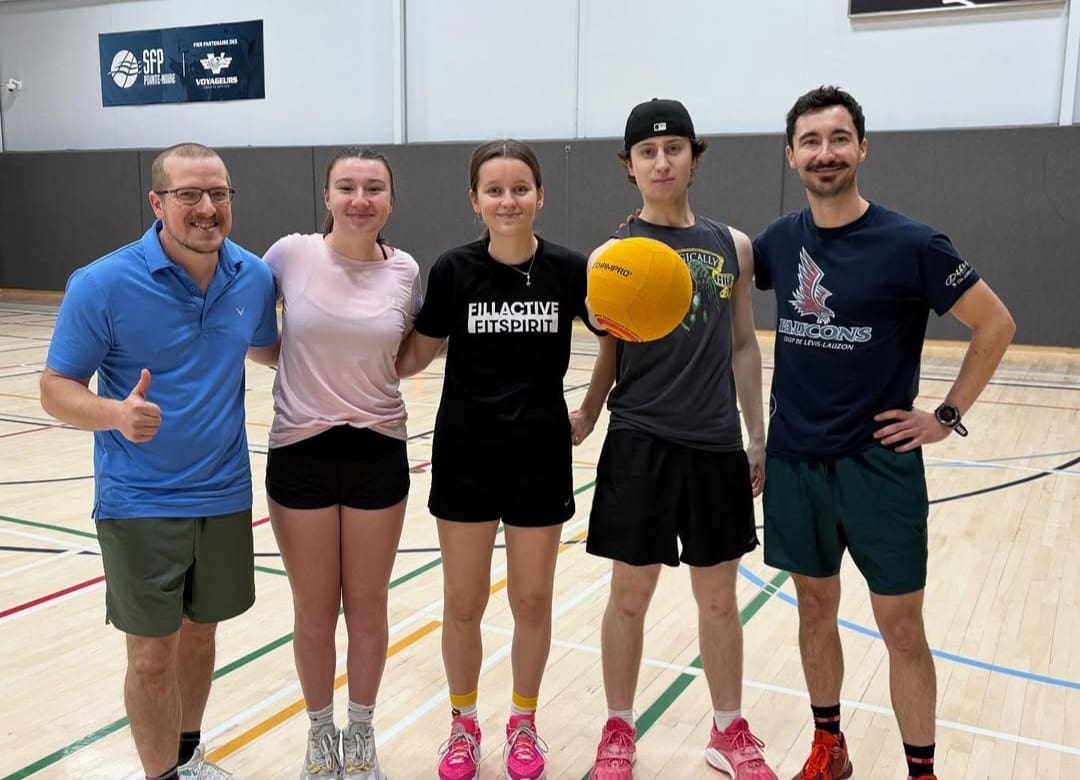 Cinq personnes se tiennent debout sur un terrain de basket dans un gymnase et sourient à la caméra. L'une d'entre elles tient un ballon jaune ; toutes sont vêtues de tenues de sport. Un homme, à gauche, lève le pouce.