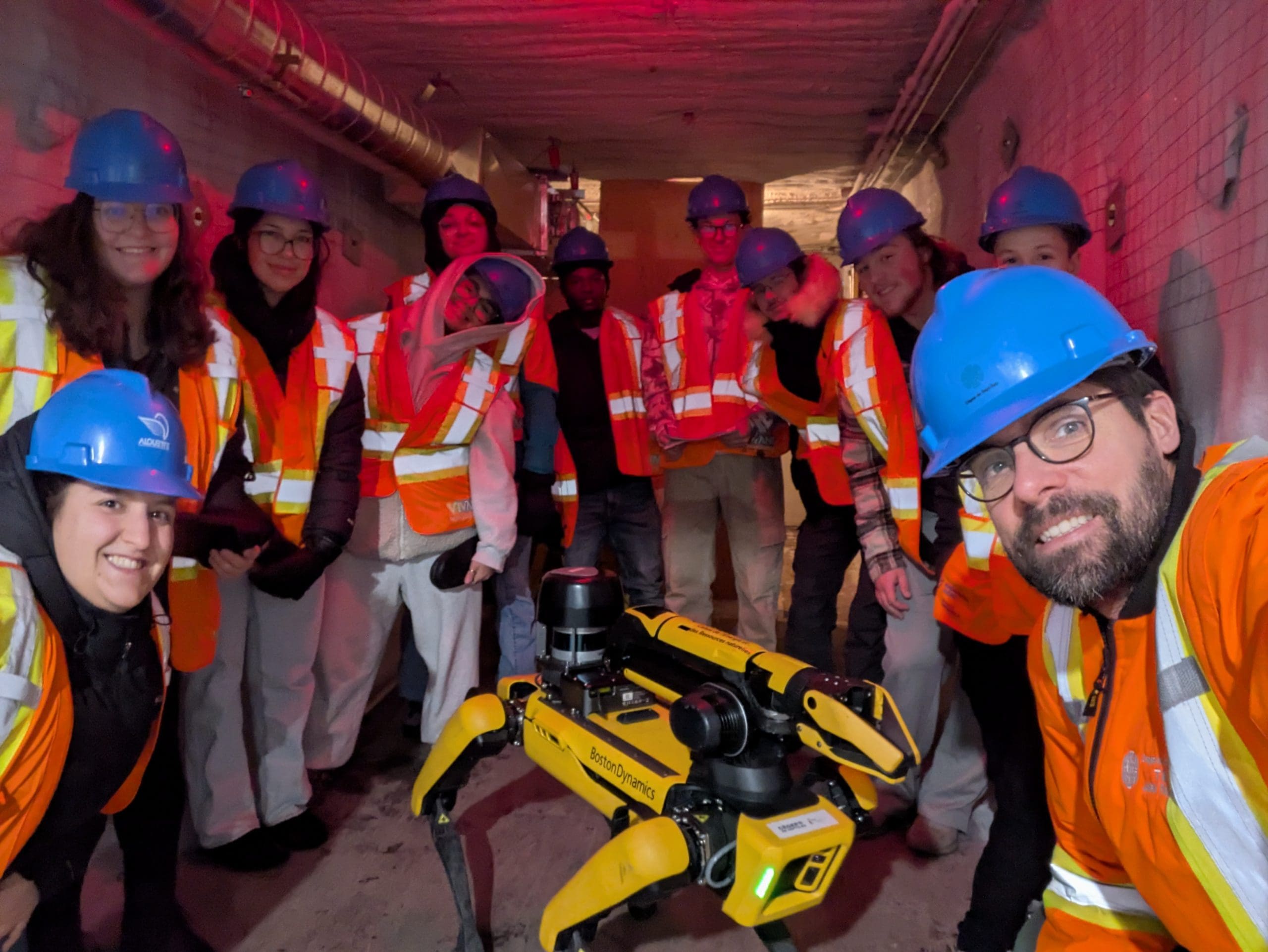 Un groupe de personnes vêtues d'équipements de sécurité et portant des casques de chantier bleus posent dans un décor industriel aux côtés d'un chien robot jaune nommé Spot.