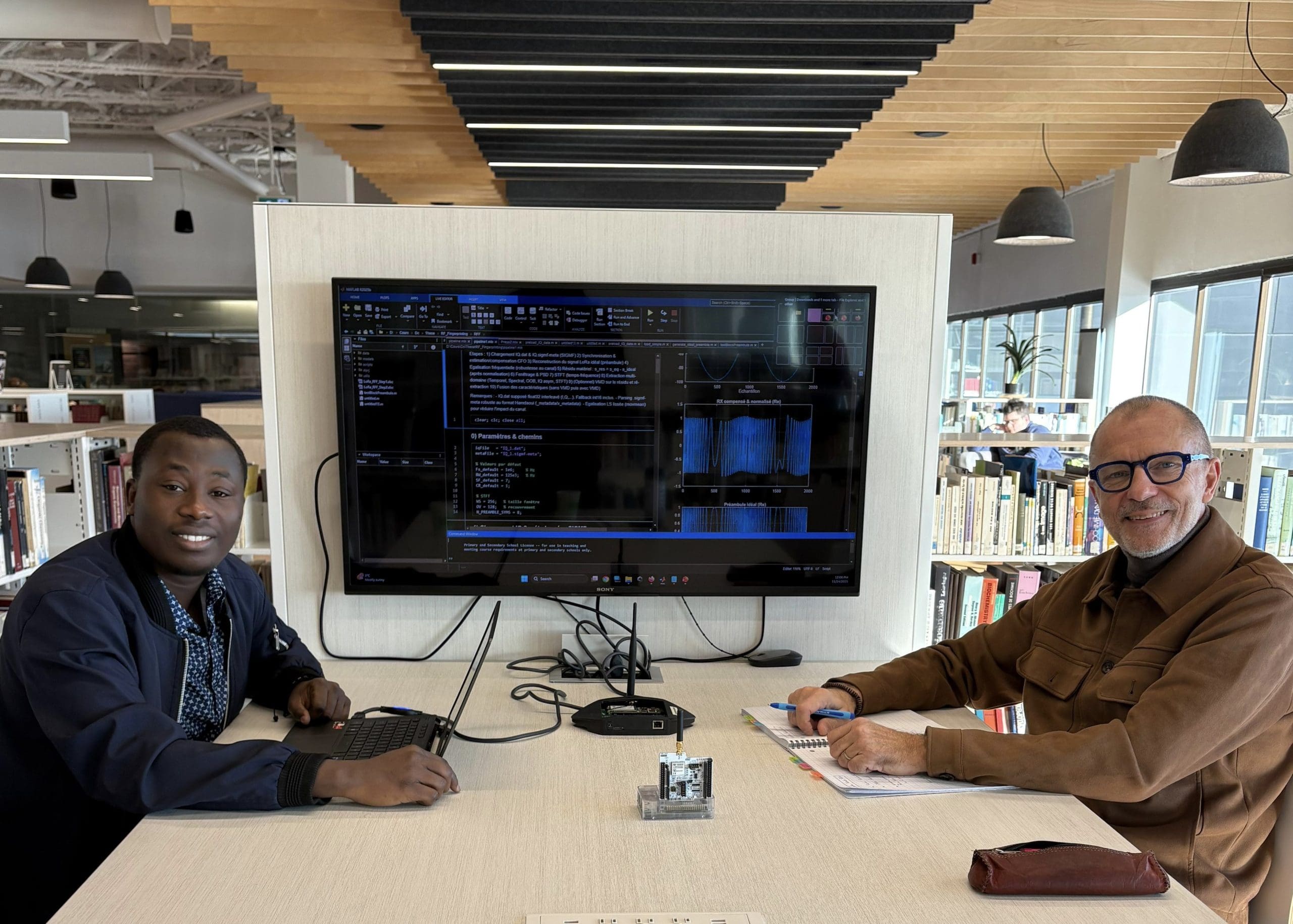 Deux hommes sont assis à une table de bibliothèque, l'un avec un ordinateur portable, l'autre avec un bloc-notes. Un grand écran, placé entre eux, affiche du code. On aperçoit des étagères et de grandes fenêtres à l'arrière-plan.