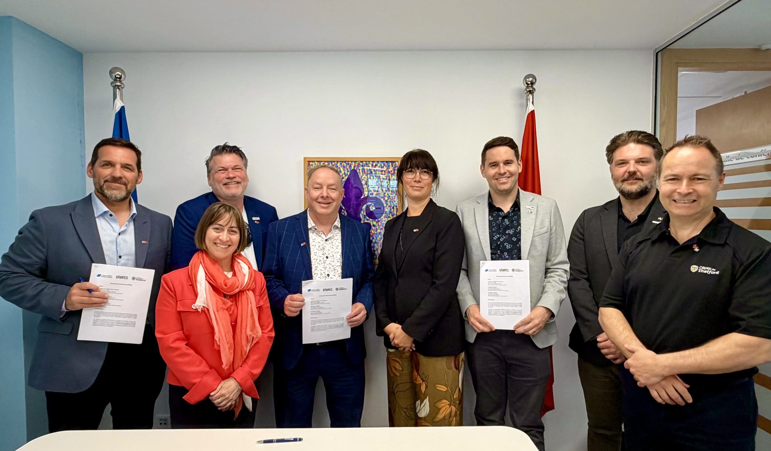 Huit personnes se tiennent à l'intérieur, souriantes et tenant des documents, devant deux drapeaux et un tableau coloré accroché au mur. Le groupe semble participer à un événement officiel ou à une cérémonie de signature.