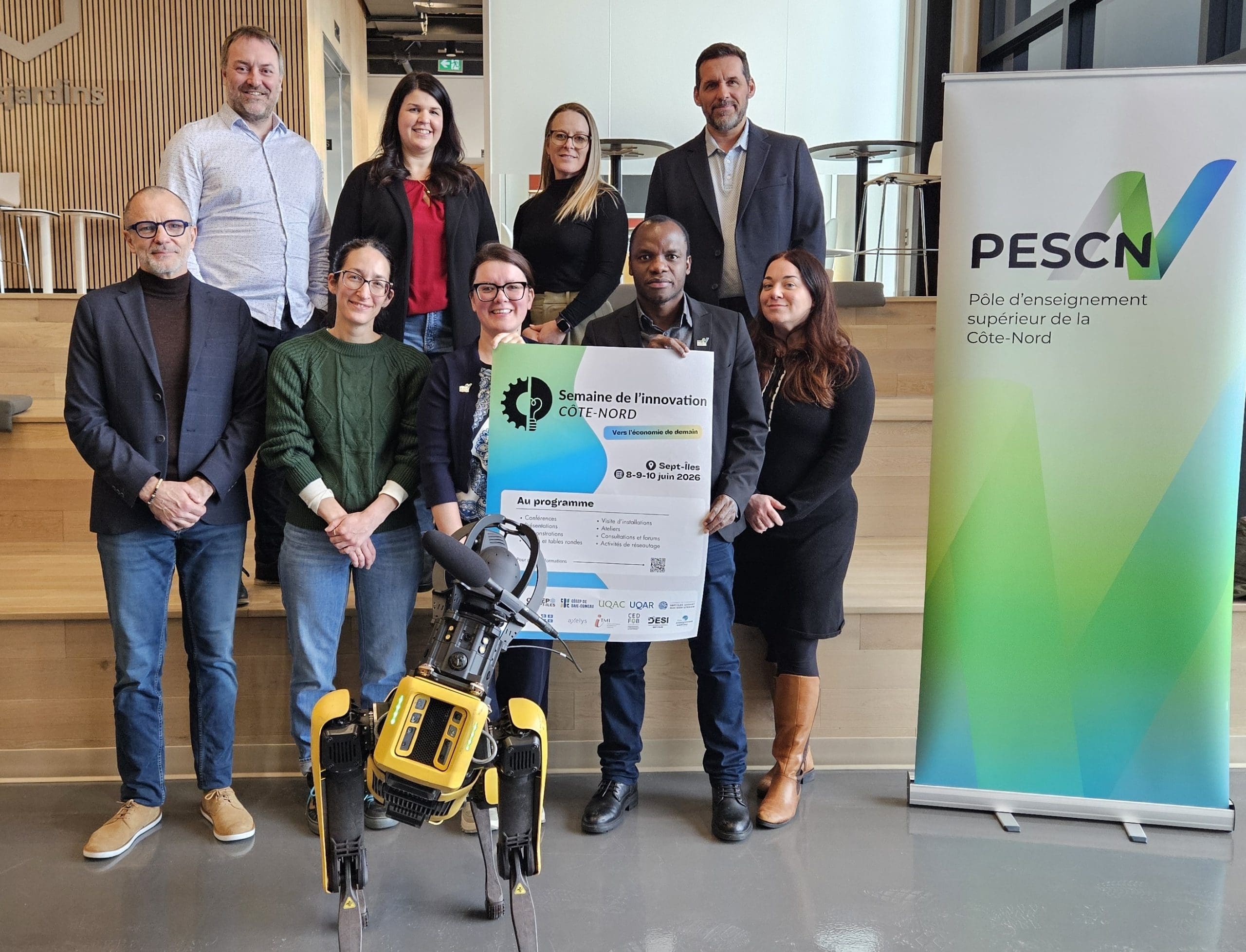 Dix personnes posent à l'intérieur avec un chien robotisé jaune et une grande affiche. Elles sont debout ou assises sur des marches en bois, à côté d'une banderole PESCN, dans un bâtiment moderne.