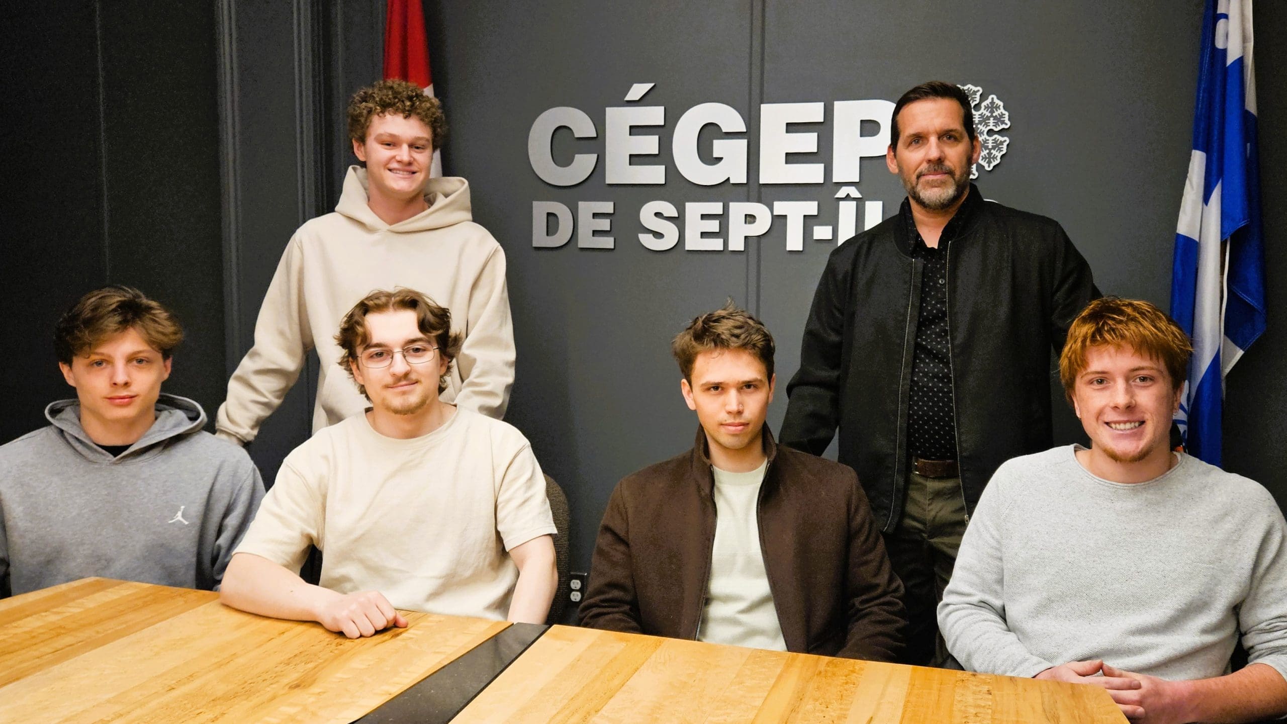 Cinq jeunes hommes et un homme plus âgé posent et sourient autour d'une table en bois, devant une pancarte murale sur laquelle on peut lire « CÉGEP DE SEPT-ÎLES », avec un drapeau canadien et un drapeau québécois visibles en arrière-plan.