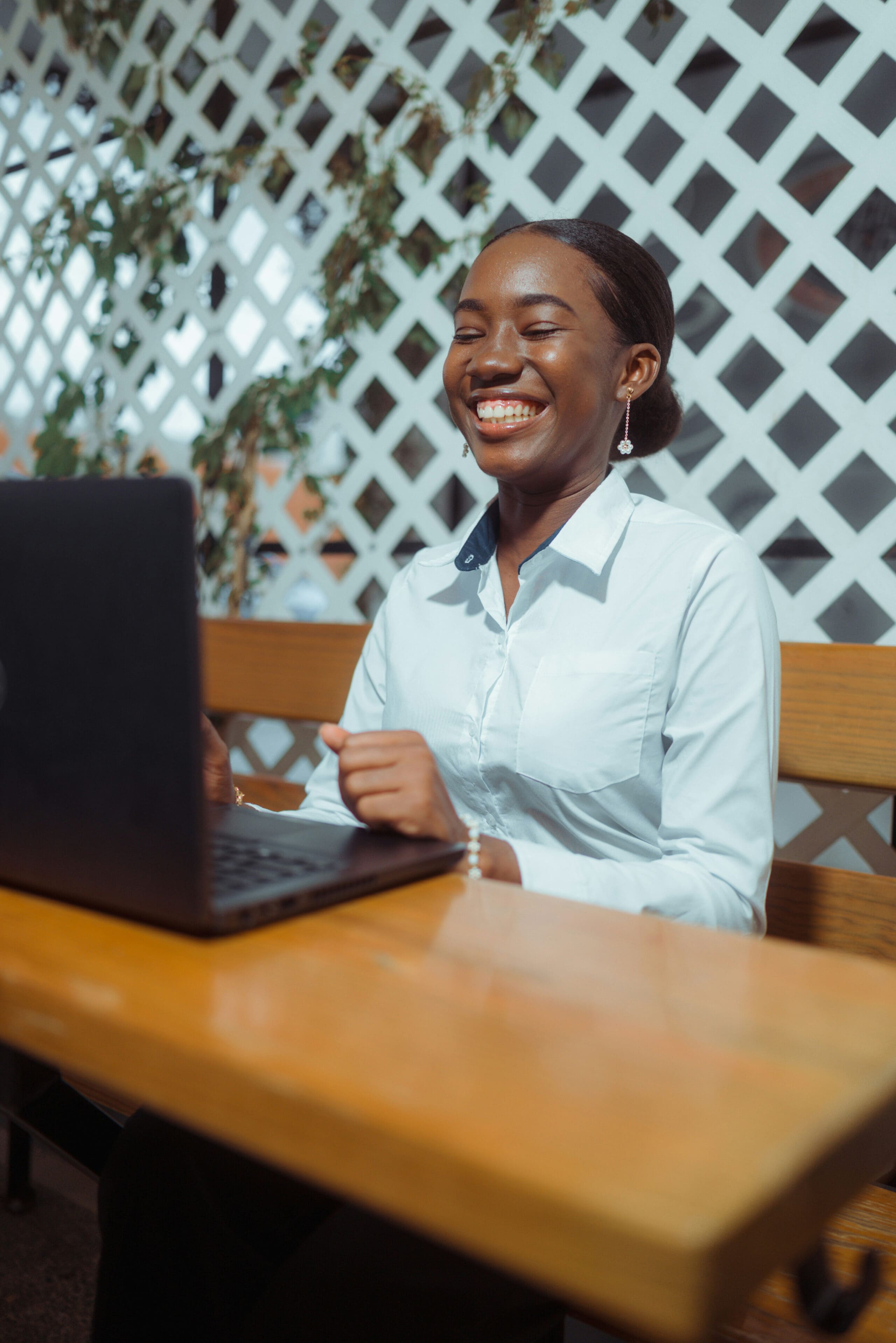 Une femme vêtue d'une chemise blanche est assise à une table en terrasse ; elle sourit et travaille sur son ordinateur portable, avec en arrière-plan une clôture en treillis blanc et des plantes vertes.