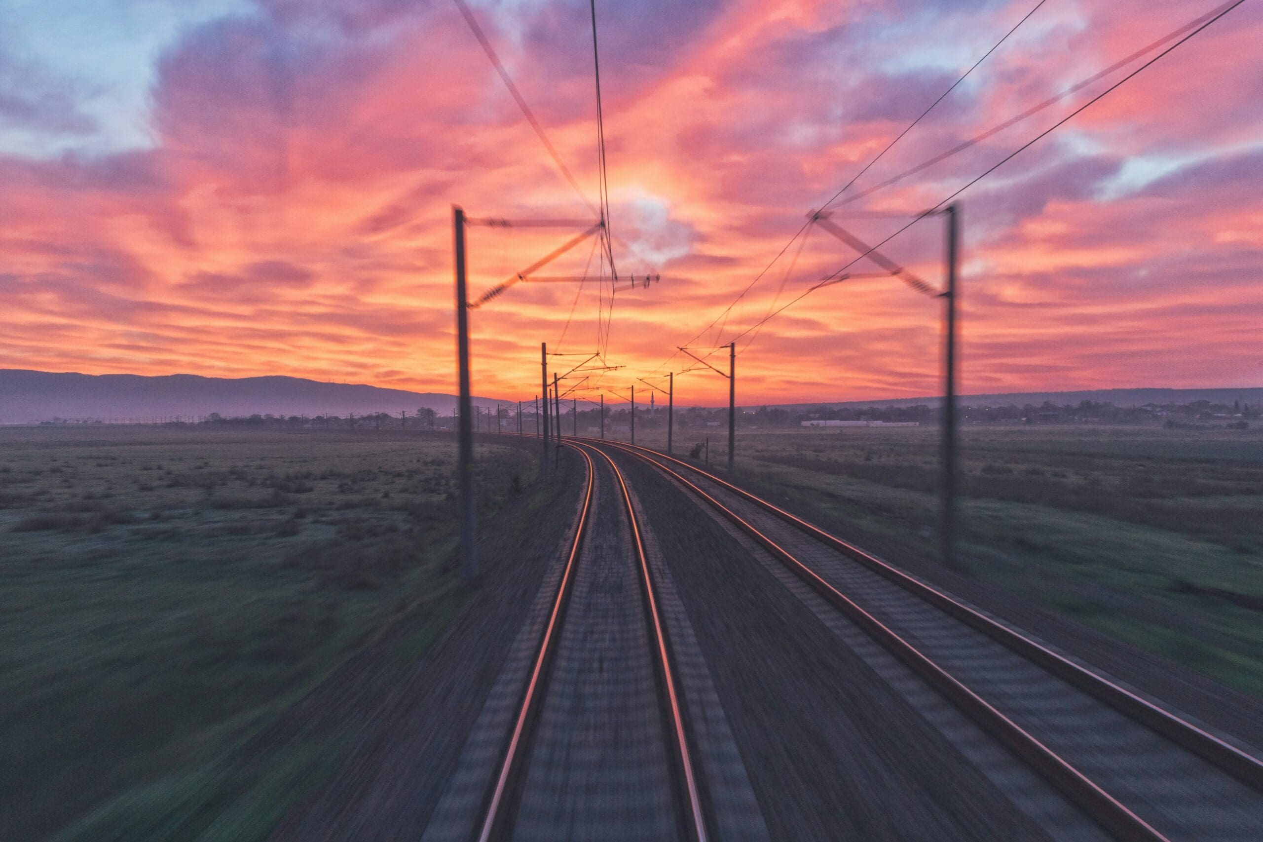 Au lever ou au coucher du soleil, les voies ferrées serpentent à travers un paysage verdoyant, sous les lignes électriques, tandis qu'un ciel spectaculaire se pare de nuages aux teintes vives de rose, de violet et d'orange.