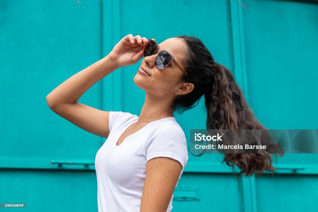 Une femme aux longs cheveux bruns attachés en queue de cheval, portant des lunettes de soleil et un t-shirt blanc, sourit et penche la tête en arrière sur un fond turquoise vif.