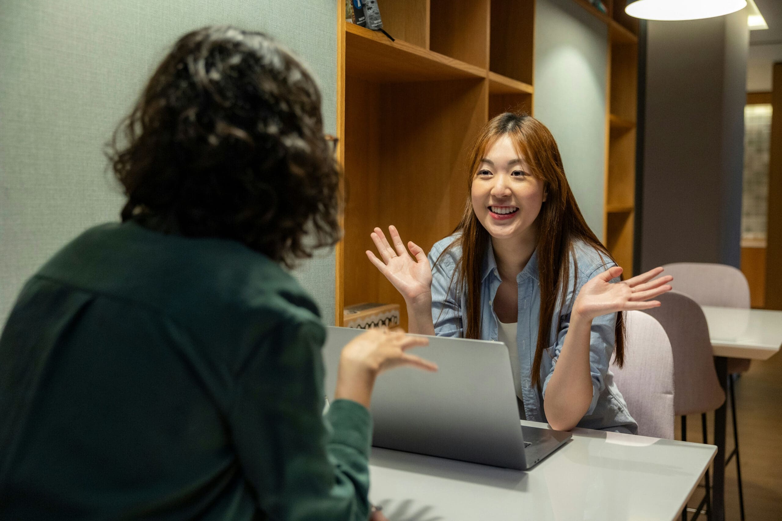 Deux femmes sont assises à une table devant un ordinateur portable ; elles discutent et sourient. L'une d'elles fait face à la caméra, les mains levées dans un élan d'enthousiasme, tandis que l'autre est vue de dos. Elles semblent avoir une conversation animée.