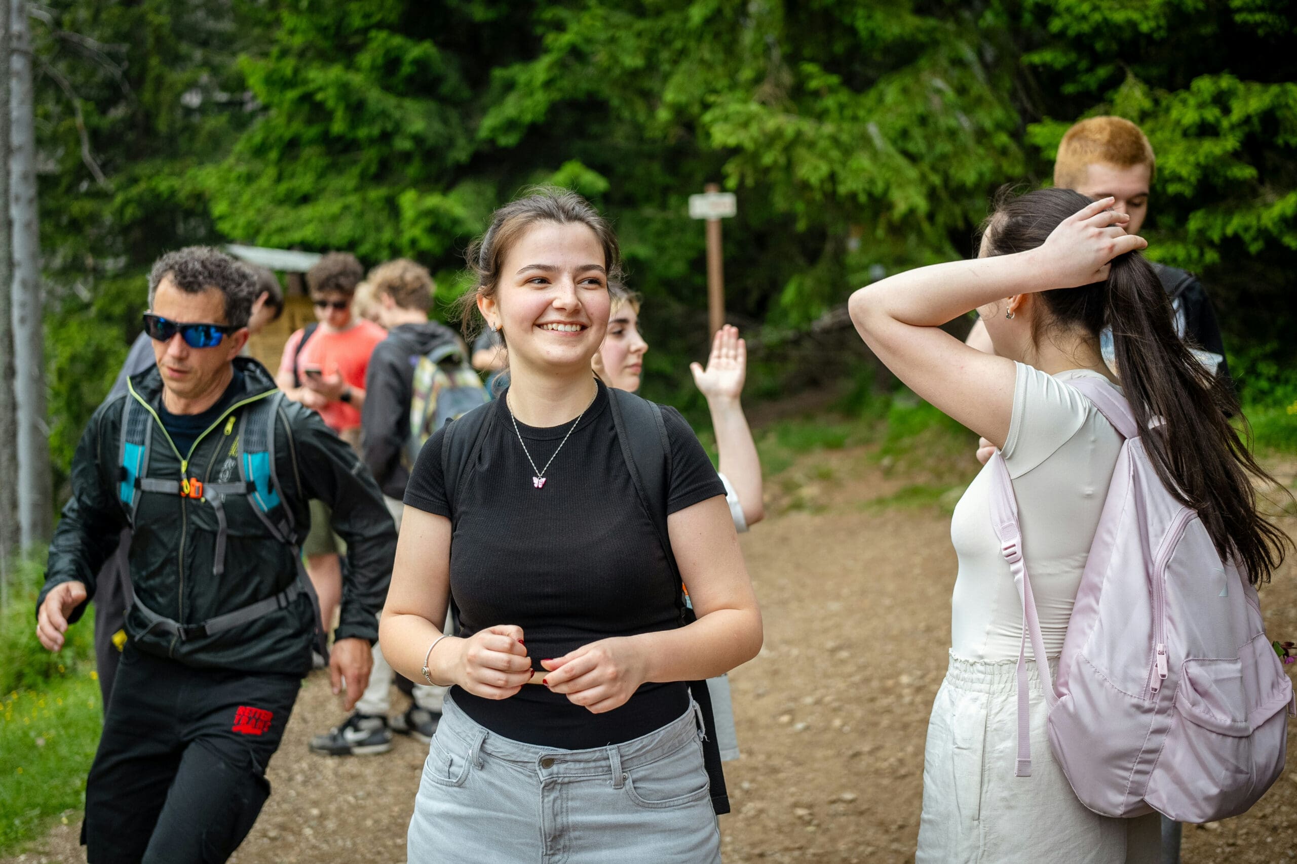 Un groupe de jeunes fait une randonnée sur un sentier forestier. Au premier plan, on aperçoit une femme souriante vêtue d'un t-shirt noir, tandis qu'à l'arrière-plan, d'autres marchent et discutent, entourés d'arbres verdoyants.