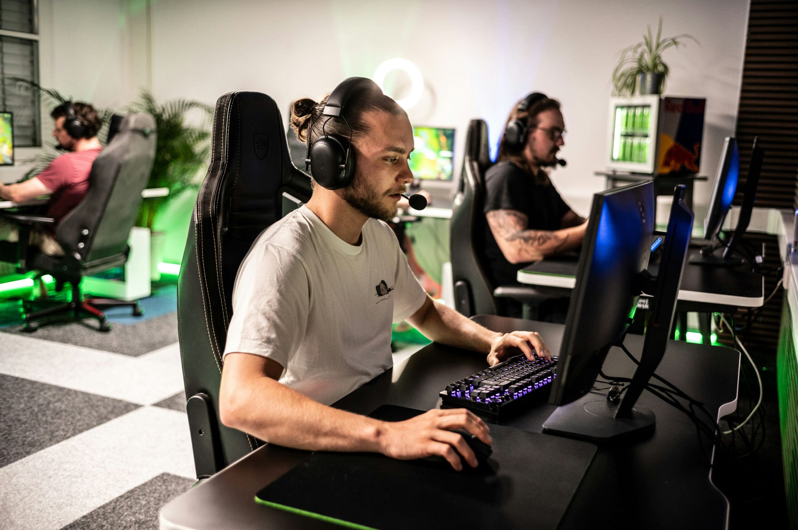 Trois personnes équipées de casques jouent à des jeux vidéo sur des ordinateurs dans une salle de jeux moderne, baignée d'un éclairage vert et agrémentée de plantes ; elles sont concentrées sur leurs écrans et utilisent un clavier et une souris.