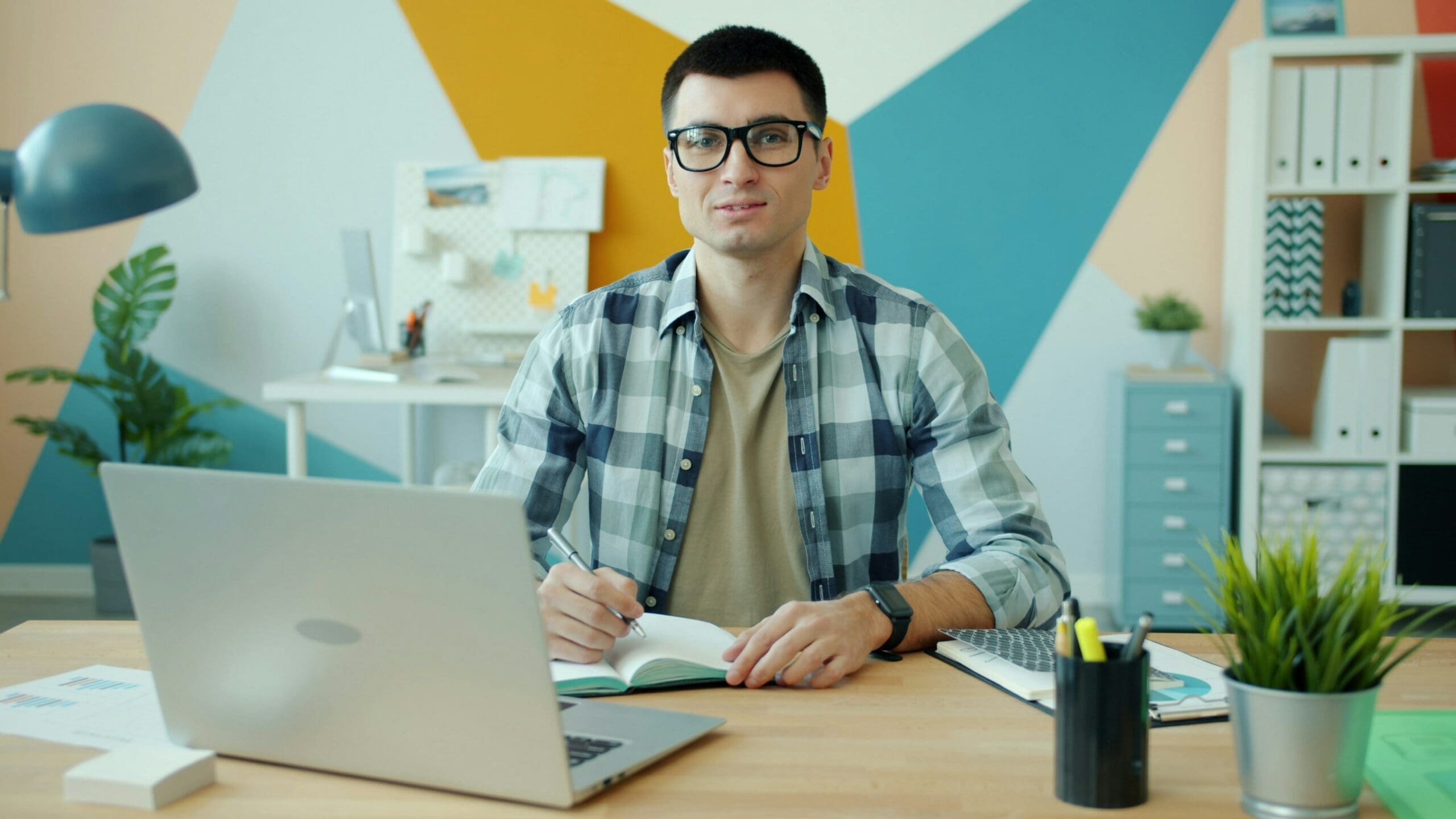 Un homme portant des lunettes et une chemise à carreaux est assis à un bureau sur lequel se trouvent un ordinateur portable, un cahier et des fournitures de bureau ; il regarde vers la caméra. En arrière-plan, on aperçoit une décoration murale géométrique colorée et des étagères de bureau.