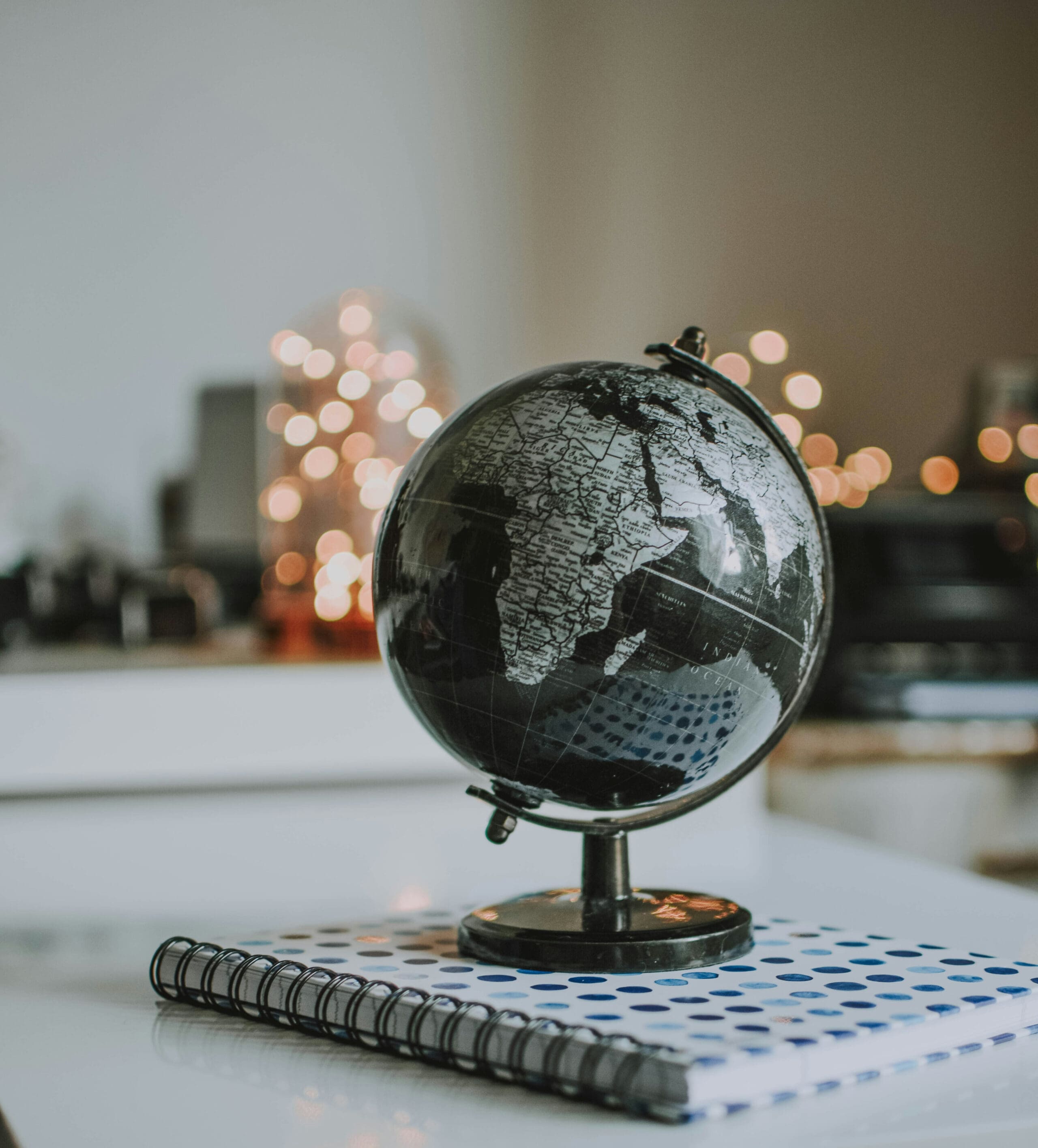 Un globe terrestre noir et argenté repose sur un cahier à spirales à pois bleus, posé sur un bureau blanc. L'arrière-plan est flou, parsemé de petites lumières chaudes et chatoyantes.
