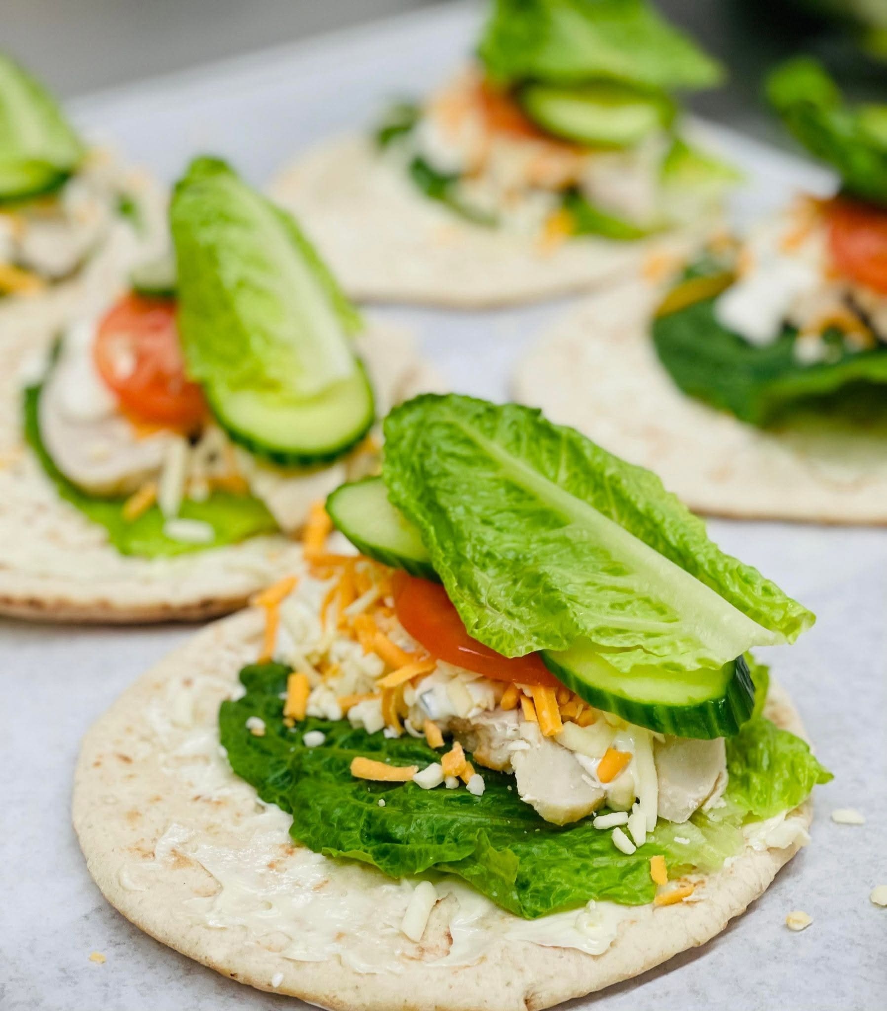 Des galettes garnies de laitue, de rondelles de concombre, de tomates, de fromage râpé et d'une tartinade onctueuse sont disposées sur un plan de travail clair, prêtes à être pliées ou servies.