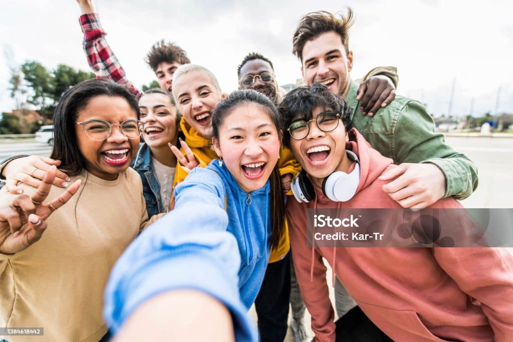 Un groupe hétéroclite de jeunes adultes souriants prend un selfie en plein air, dans la bonne humeur, en se serrant les uns contre les autres et en faisant le signe de la paix par une journée nuageuse.