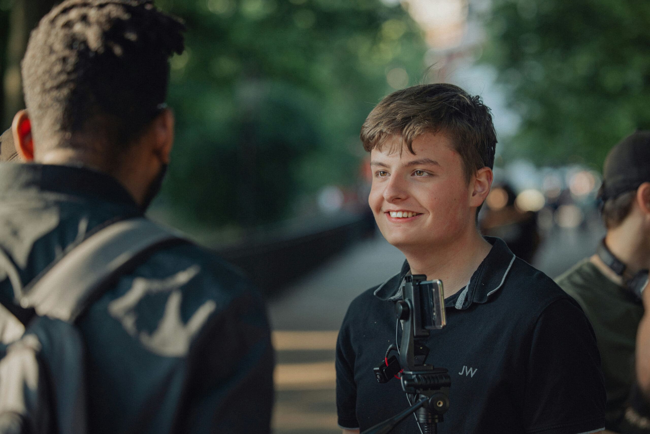 Un jeune homme muni d'un trépied pour appareil photo sourit tout en discutant avec une autre personne à l'extérieur, avec des arbres et la lumière du soleil en arrière-plan.