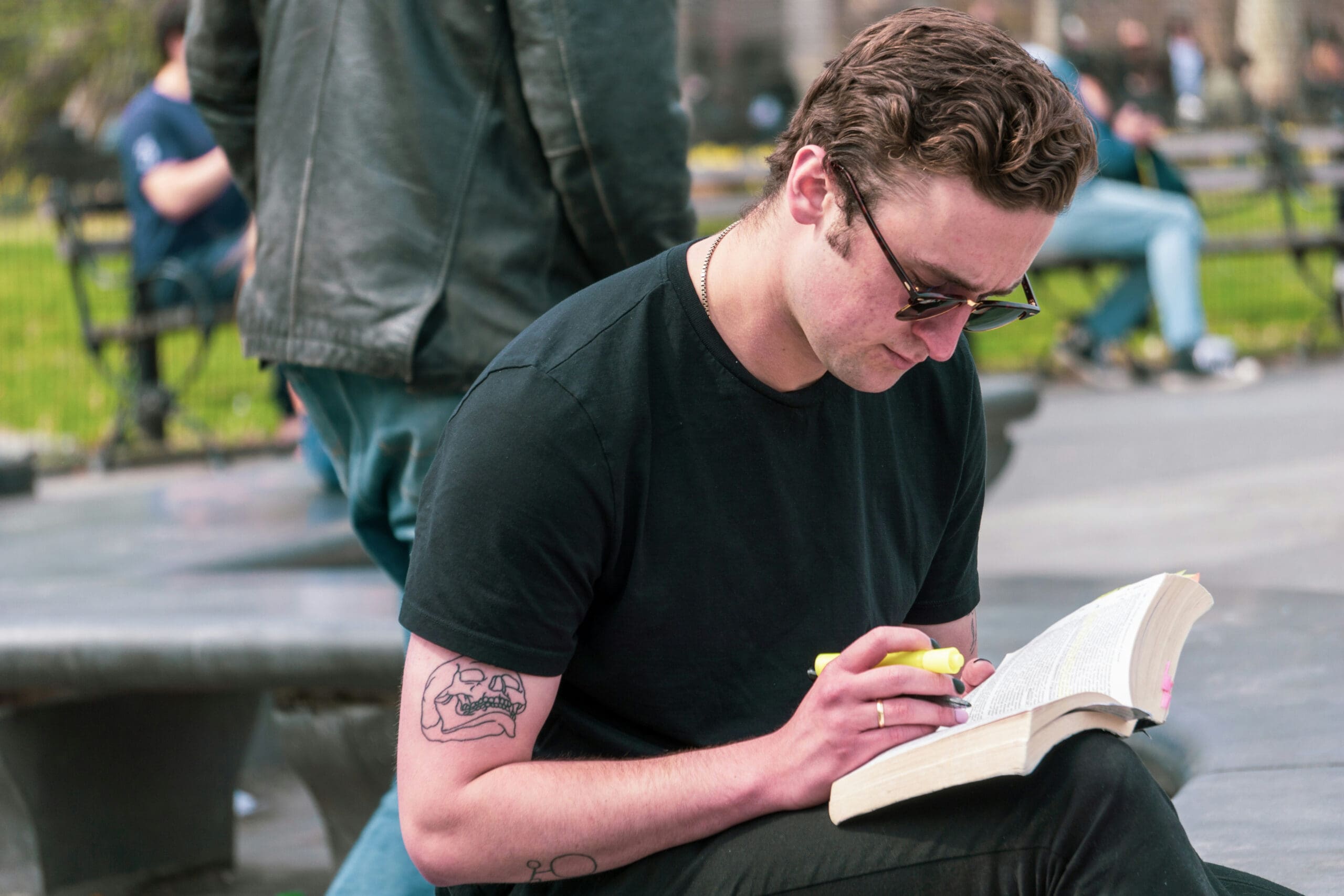 Une personne portant des lunettes de soleil et un t-shirt noir est assise à l'extérieur, en train de surligner un passage dans un livre ouvert. On aperçoit un tatouage sur son bras. D'autres personnes et de la végétation apparaissent à l'arrière-plan.