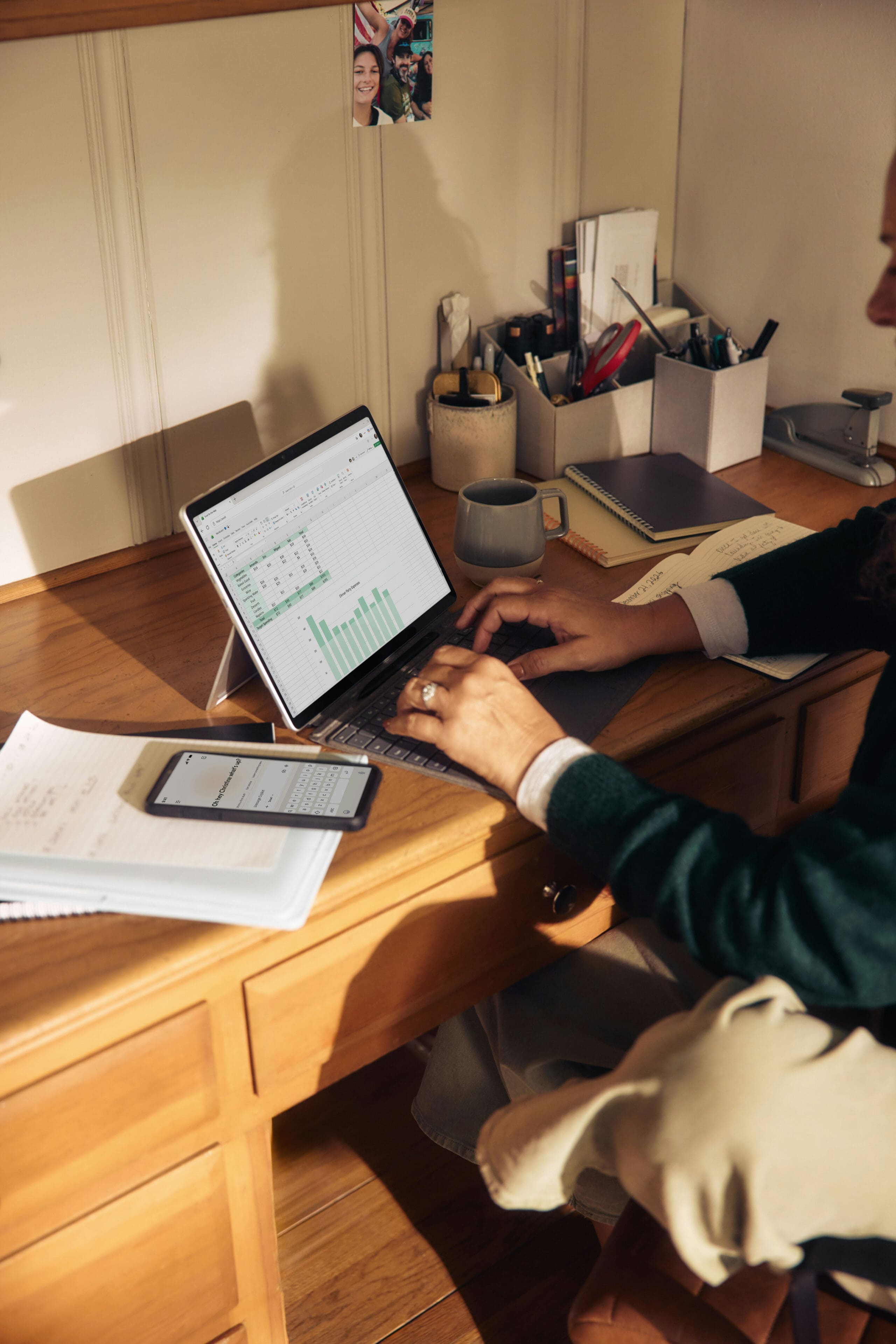 Personne travaillant à un bureau en bois, devant un ordinateur portable affichant un graphique à barres, un smartphone, des papiers, un cahier, une tasse et divers articles de bureau en arrière-plan. Seules ses mains et une partie de son corps sont visibles.