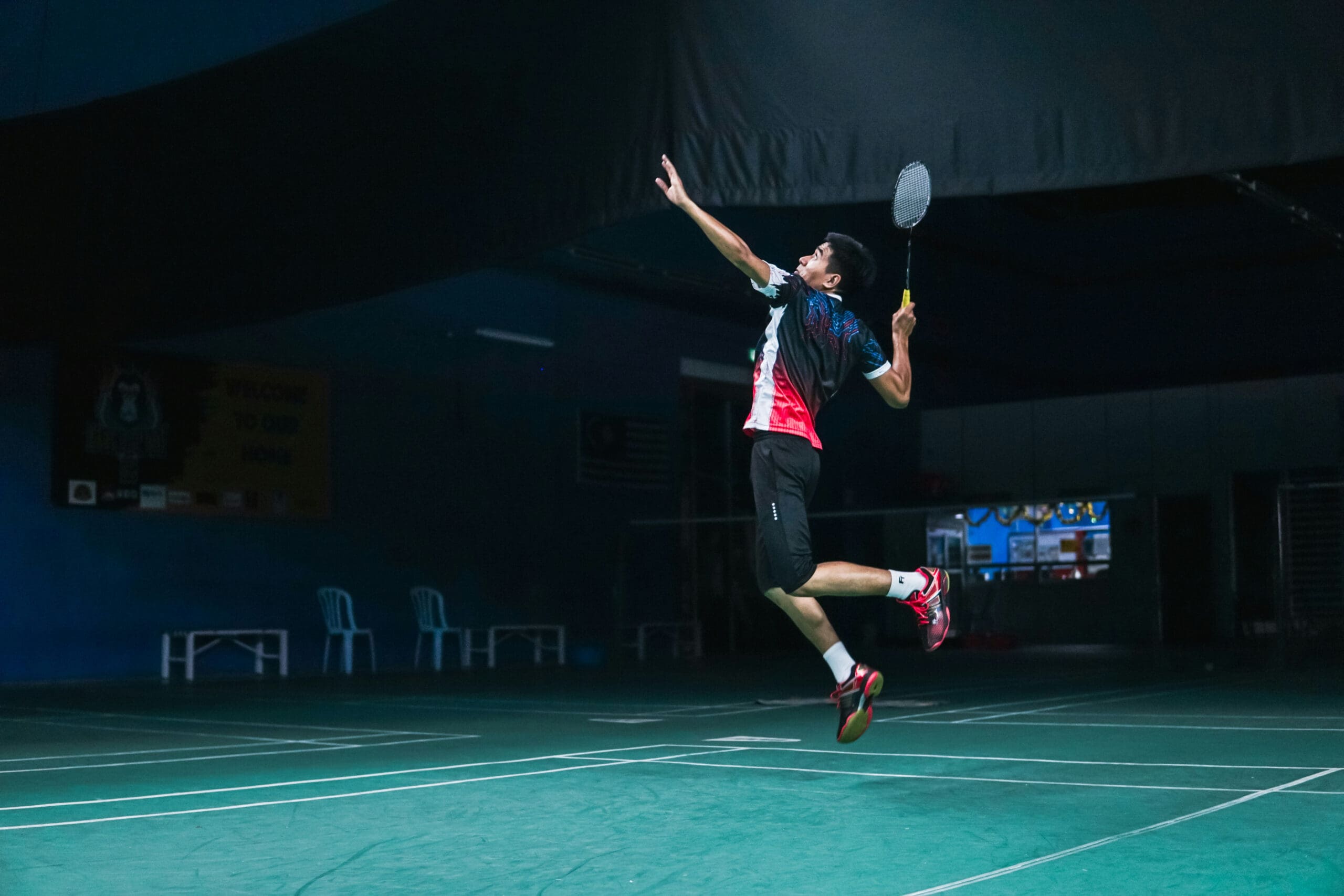 Un joueur de badminton, en plein vol, s'apprête à frapper un volant avec sa raquette sur un terrain couvert. La scène est faiblement éclairée et le joueur porte une tenue de sport ainsi que des chaussures rouges.