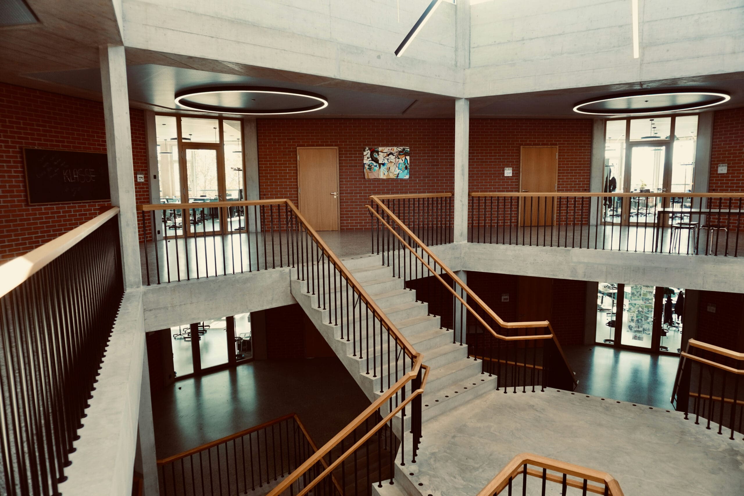 Un escalier intérieur moderne, doté d'une rampe en bois et de marches en béton, entouré de murs en briques, de grandes fenêtres et de plafonniers ronds, dans un intérieur spacieux et bien éclairé.