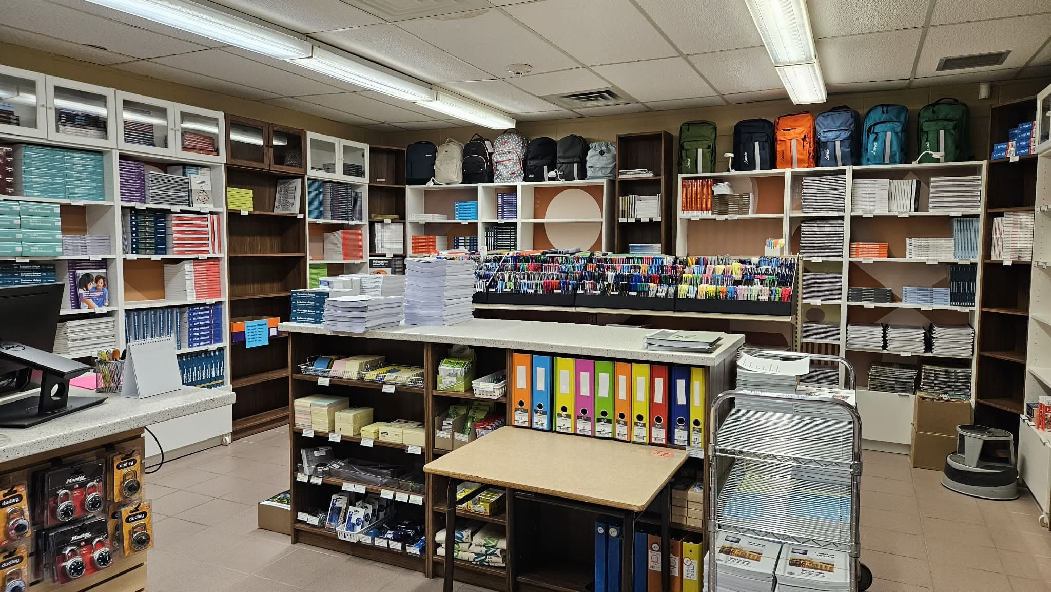 Une librairie ou une papeterie bien organisée, avec des rayons remplis de livres, de classeurs, de sacs à dos, de cahiers, de stylos et de fournitures de bureau, le tout soigneusement disposé autour d'un comptoir et de tables.